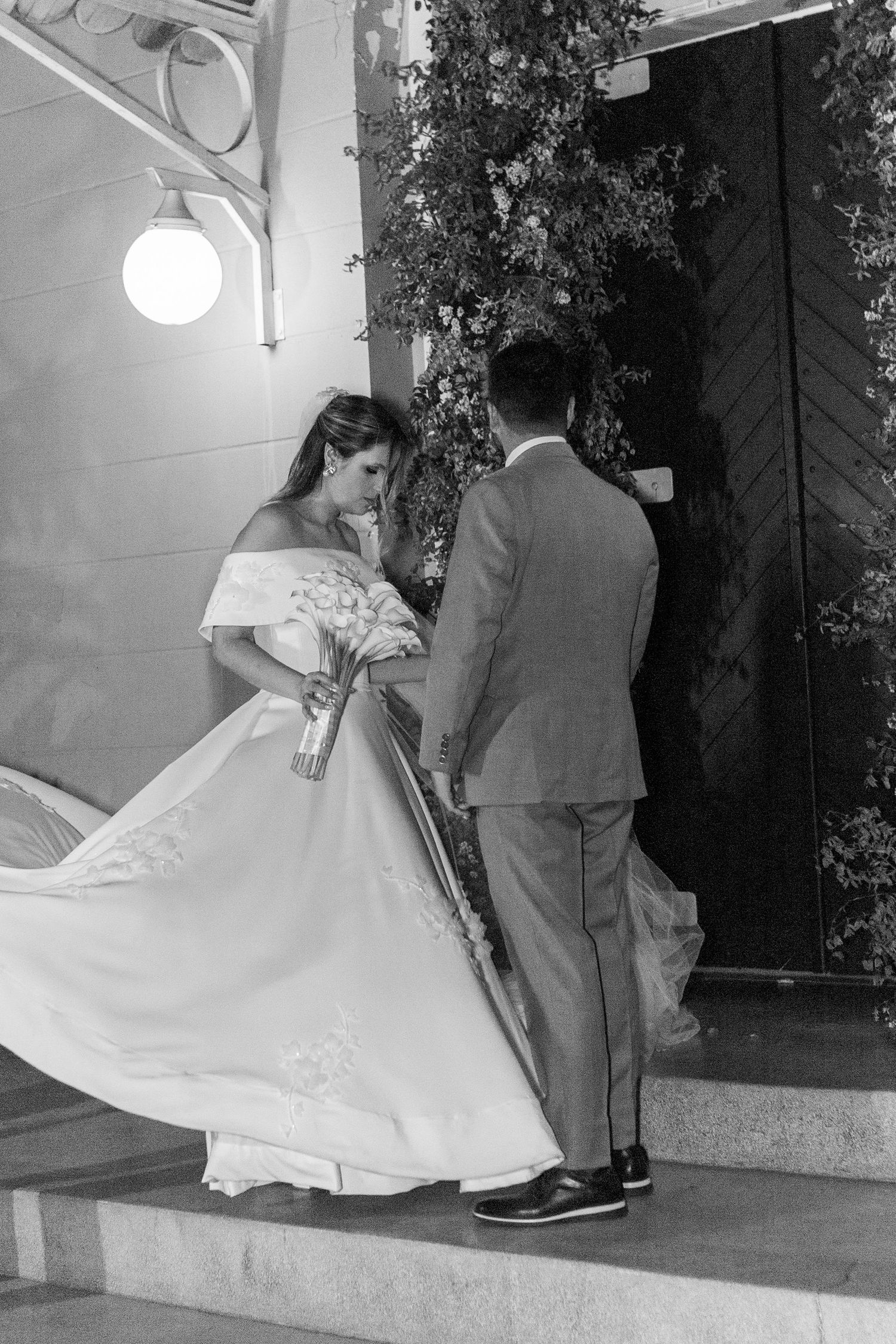 Black and white photo of a bride in a flowing gown and groom in a suit ascending steps at a wedding venue.