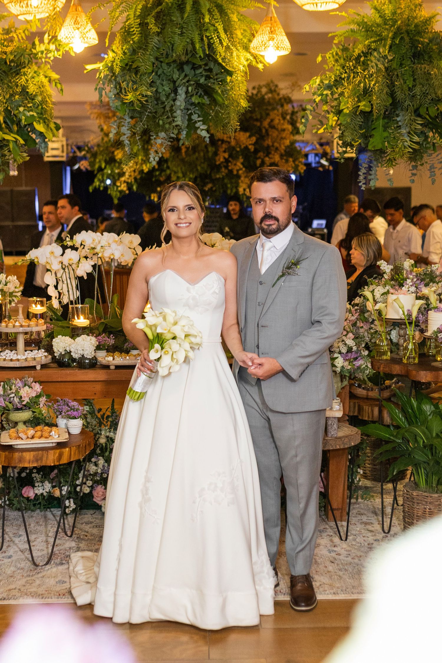 Bride in white gown holding bouquet stands with groom in gray suit at elegant floral wedding reception.