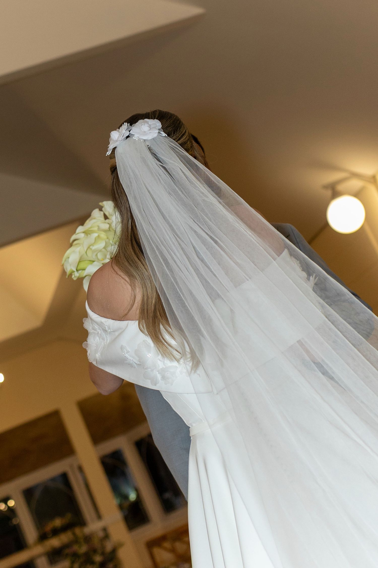 Bride in white wedding dress and veil holding bouquet, photographed from behind in elegant indoor setting.