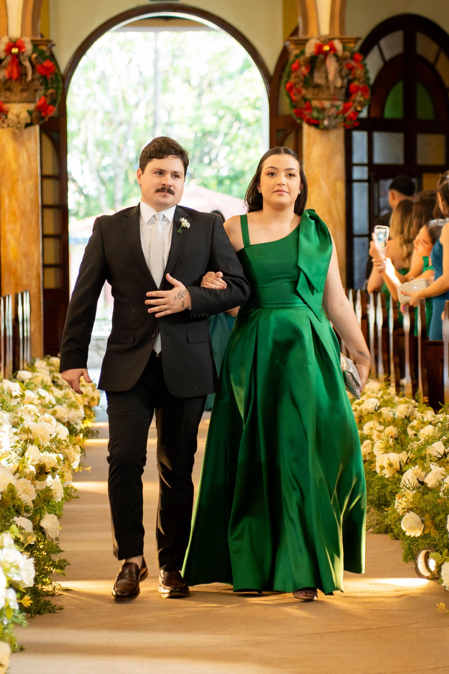 Couple walking down a floral-decorated church aisle, man in black suit, woman in elegant green gown.