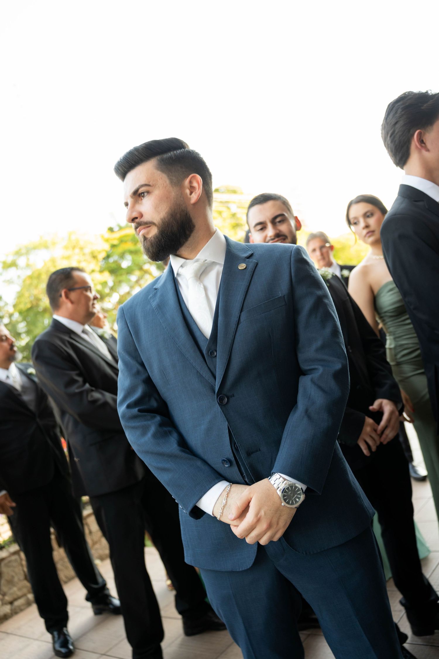 Groom in navy blue three-piece suit with white tie and watch stands confidently at outdoor wedding ceremony.