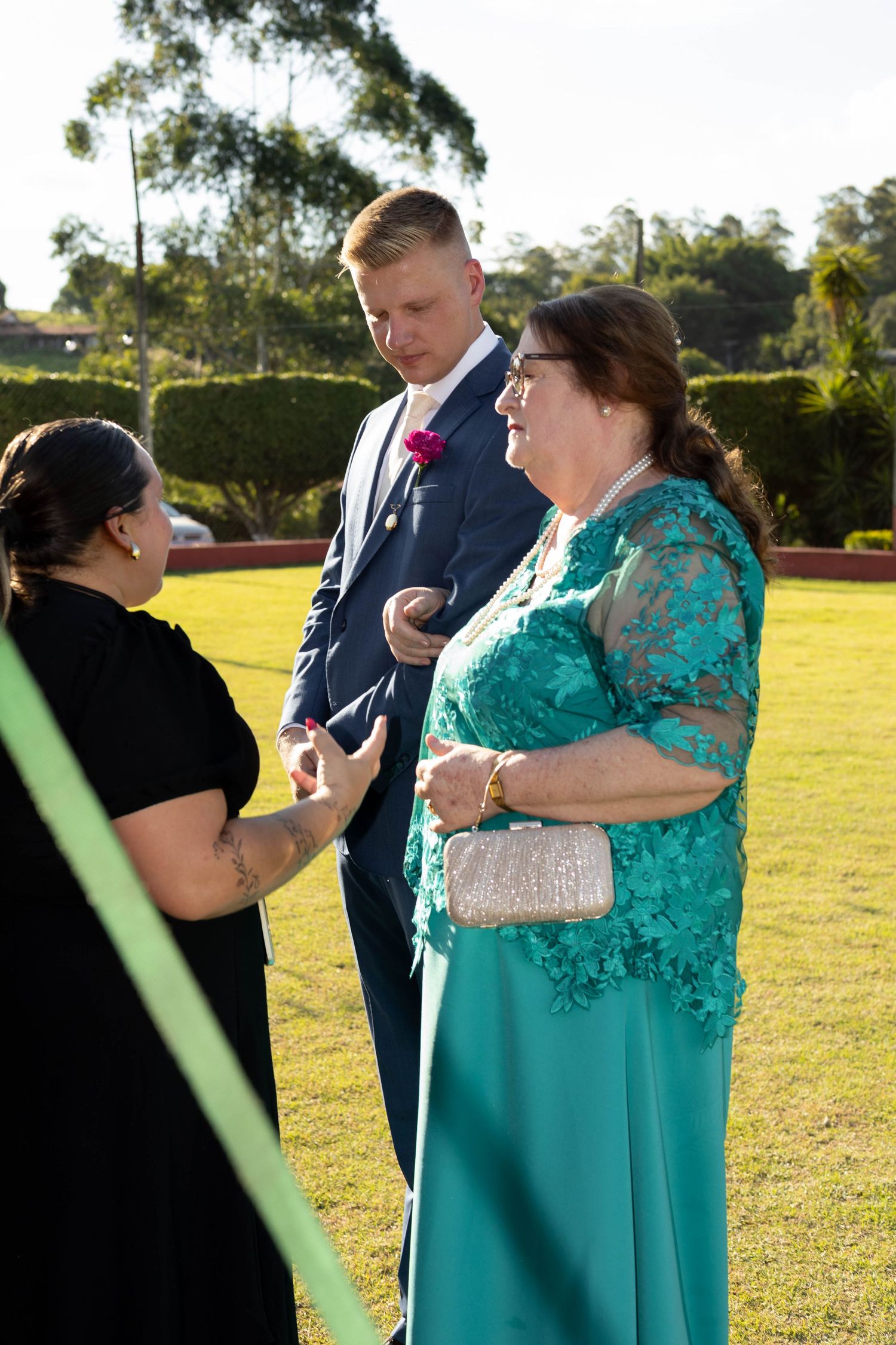 Man in blue suit and woman in teal lace gown converse with guest outdoors at a formal garden event.