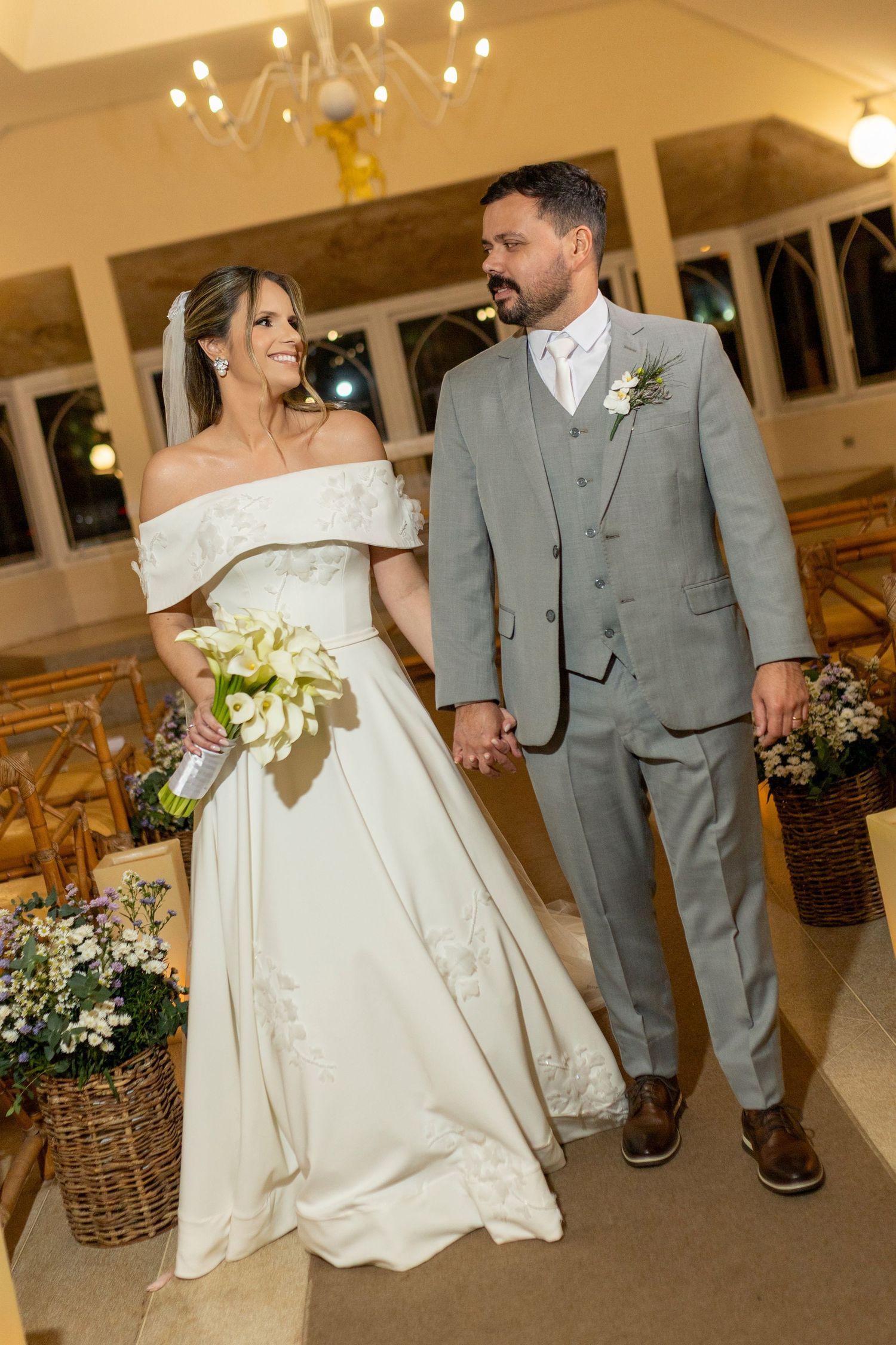 Bride in off-shoulder white gown holding bouquet walks with groom in gray suit down church aisle.