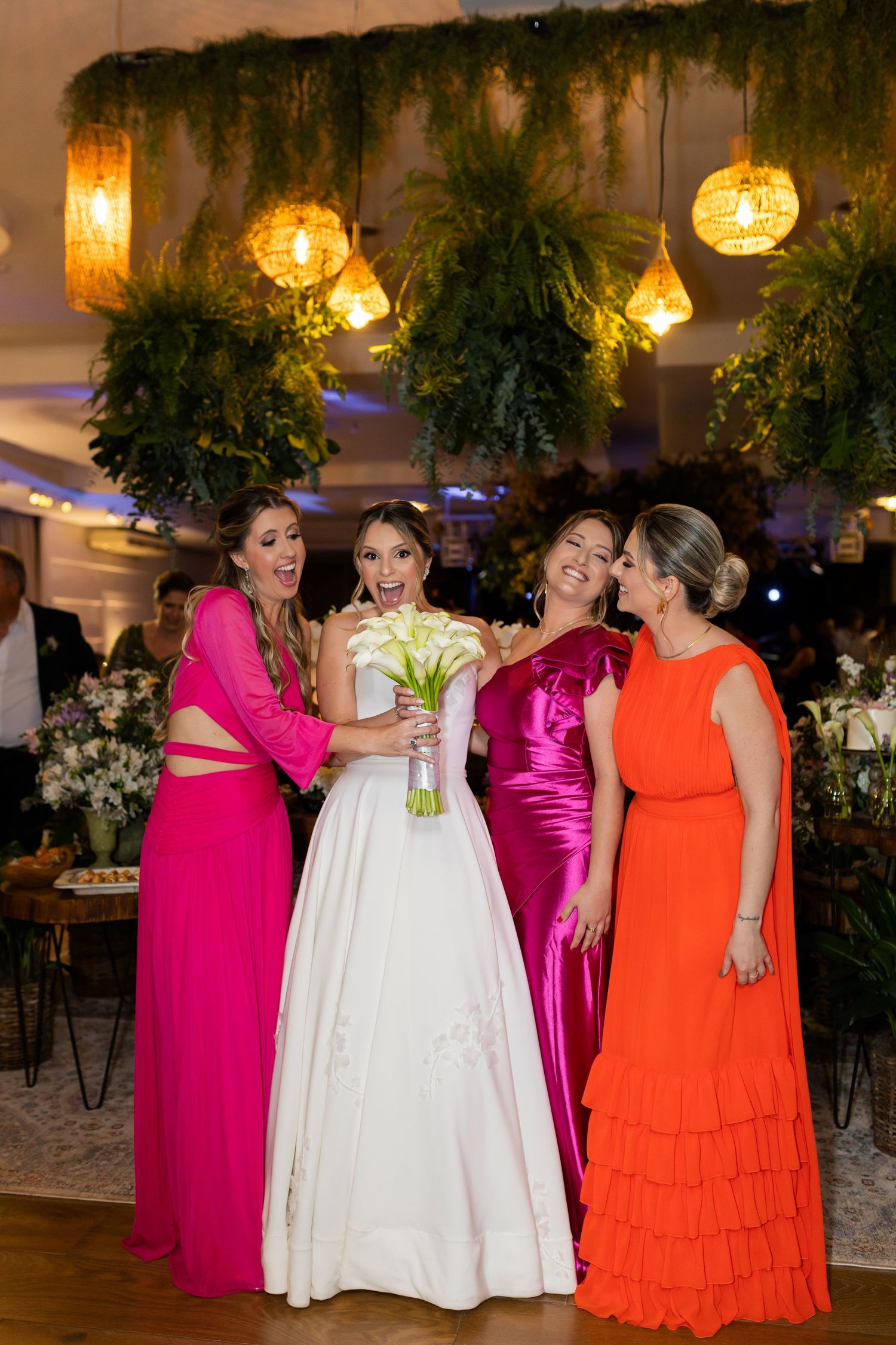 Bride in white gown holds bouquet with three women in vibrant pink and orange dresses laughing at a wedding reception.