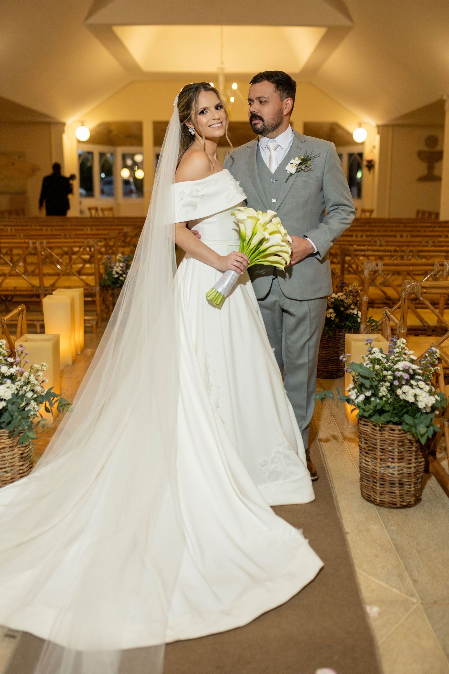 Bride and groom posing together in a warmly lit church aisle, bride holding white calla lily bouquet with long veil.