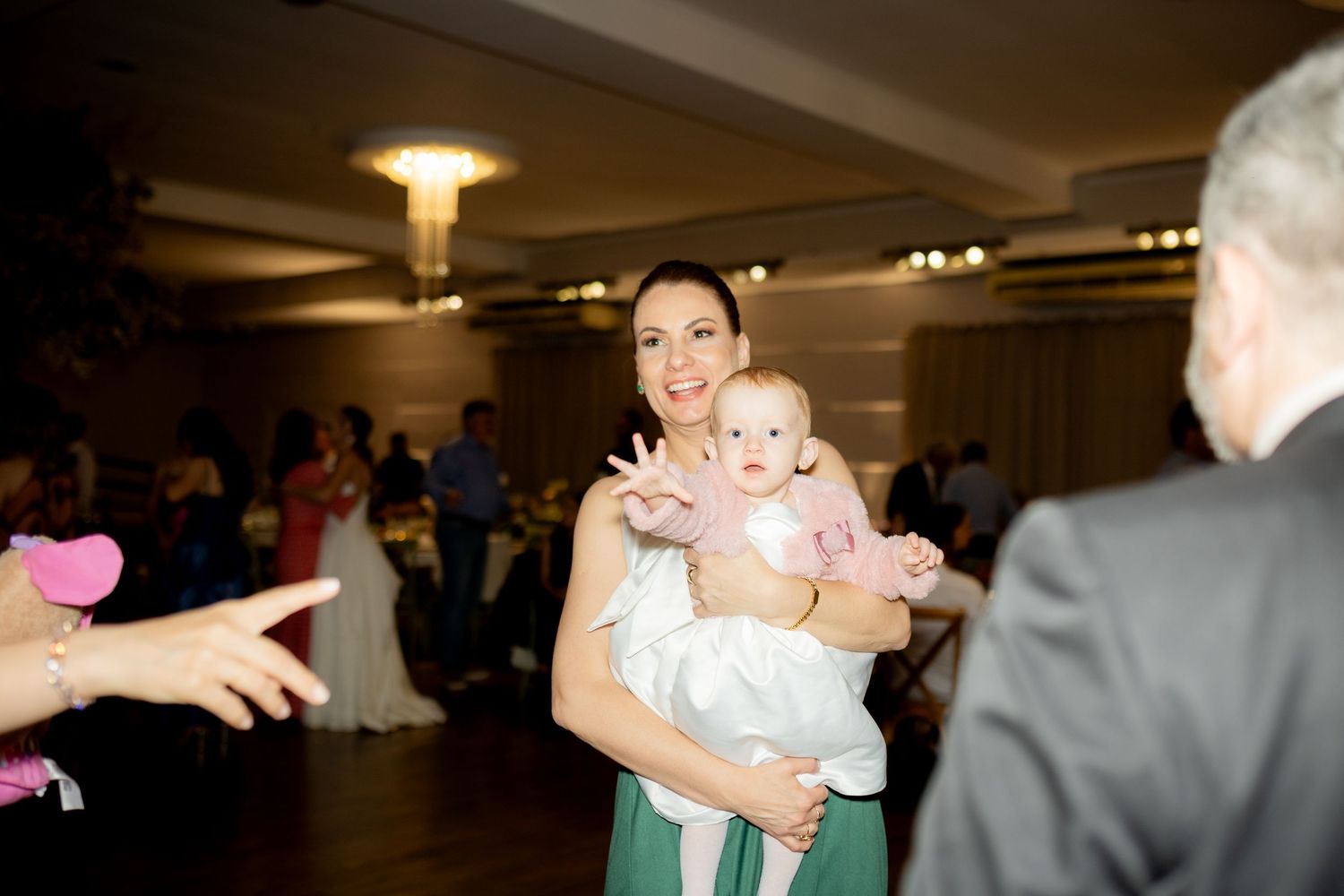 Woman holding a baby at an elegant indoor event, smiling as guests celebrate in a warmly lit banquet hall.