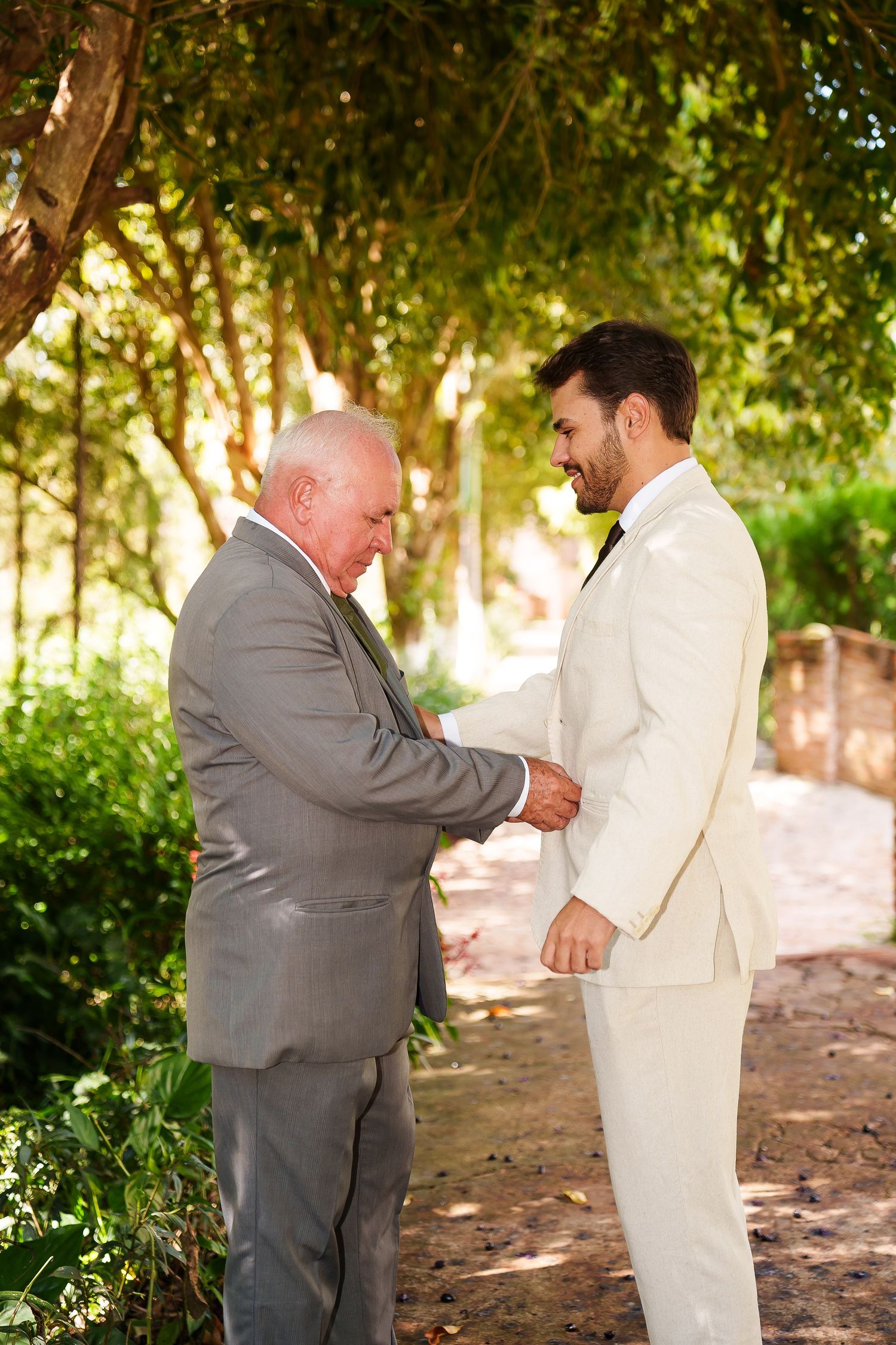Groom in white suit shaking hands with older man in gray suit outdoors under green trees at a wedding.