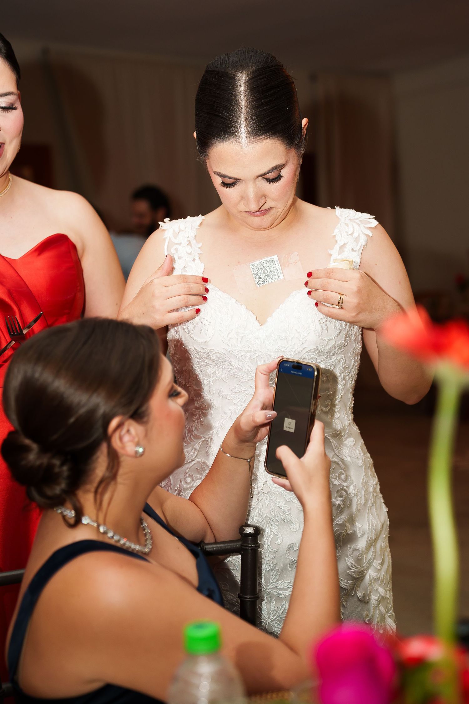 Bride in white lace gown has phone scanned at wedding reception while bridesmaid in red assists.