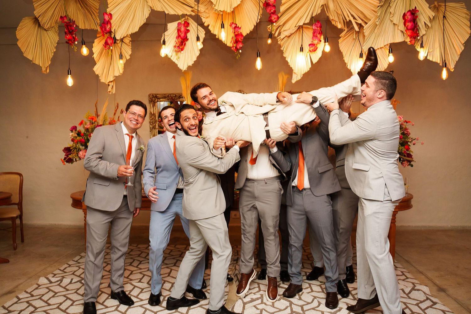 Groomsmen in gray suits lift groom overhead, laughing joyfully beneath floral ceiling decor at a festive wedding celebration.