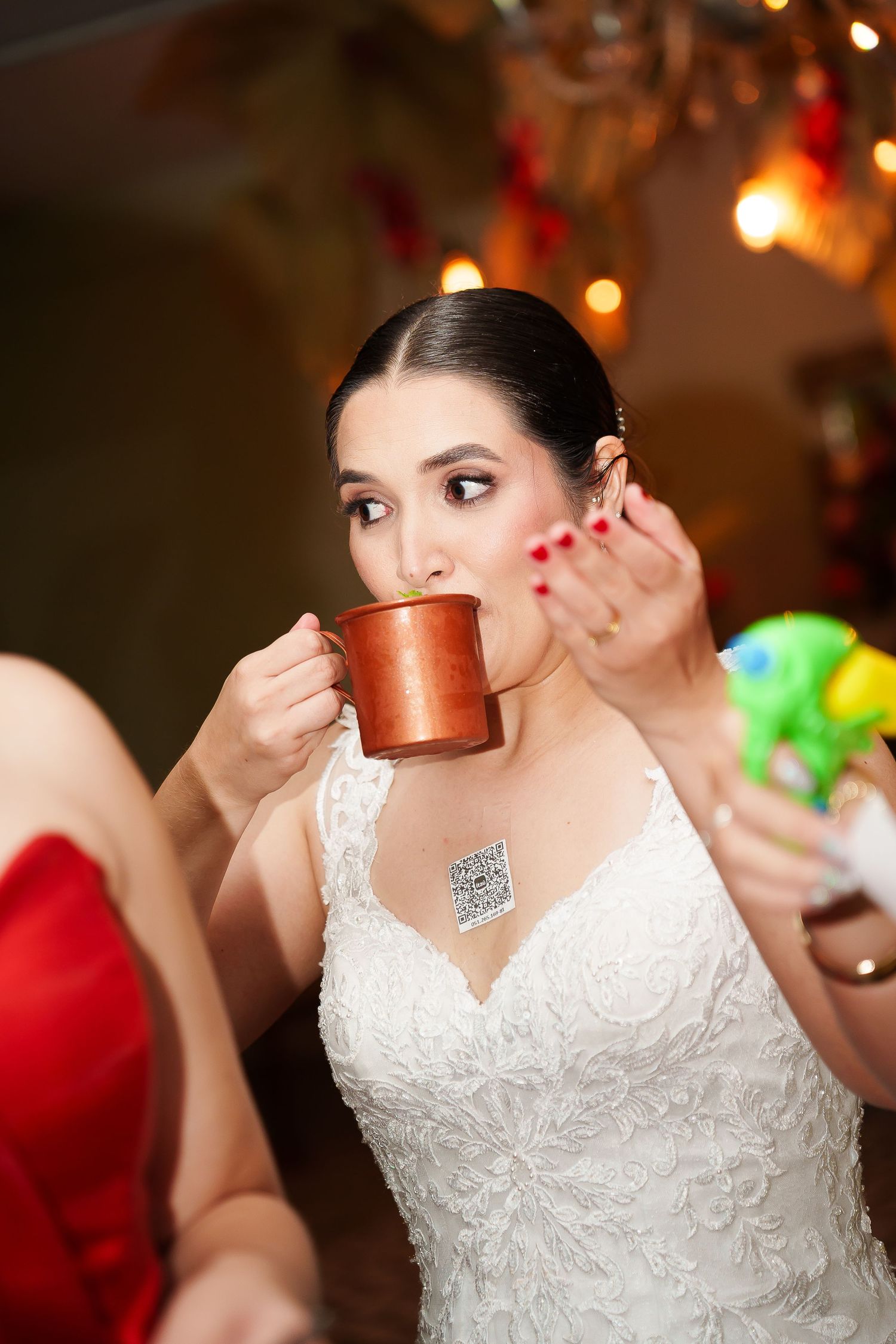 Bride in white lace gown drinking from copper mug at festive celebration with bokeh lights in background.