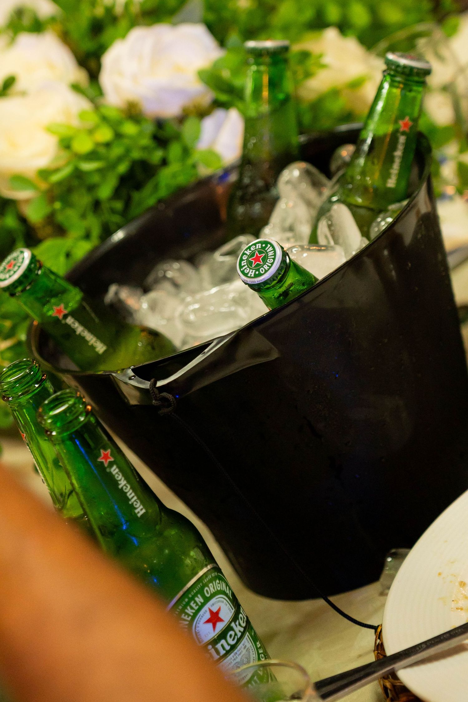 Heineken beer bottles chilling in a black ice bucket at an outdoor event with green foliage in the background.
