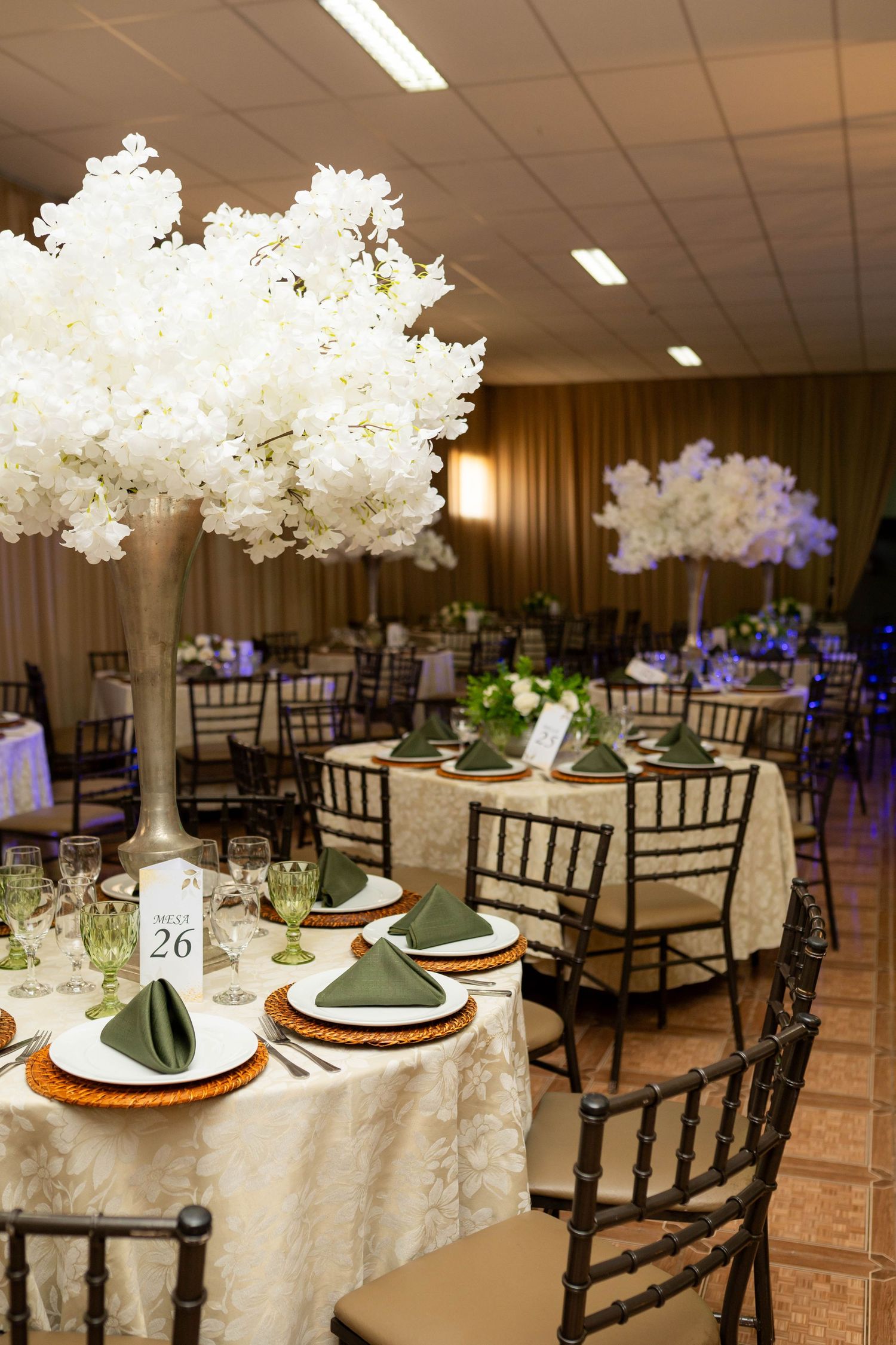 Elegant wedding reception hall with tall white floral centerpieces, green napkins, gold chargers, and dark Chiavari chairs at table 26.