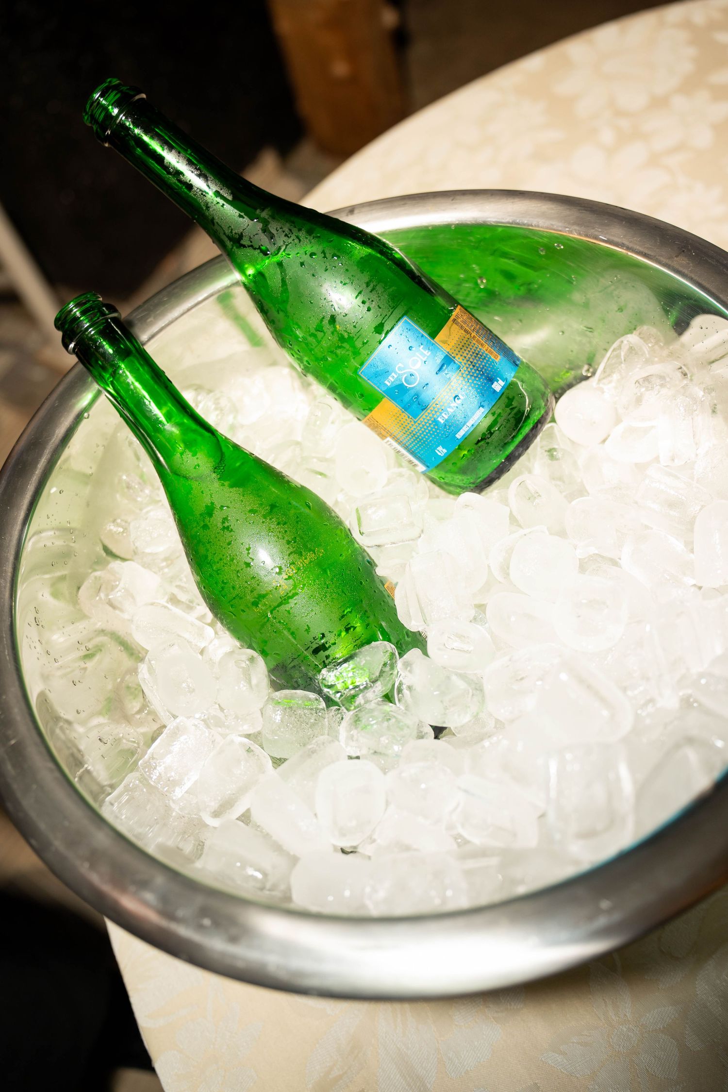 Two green glass bottles chilling in a metal ice bucket filled with ice cubes at an event.