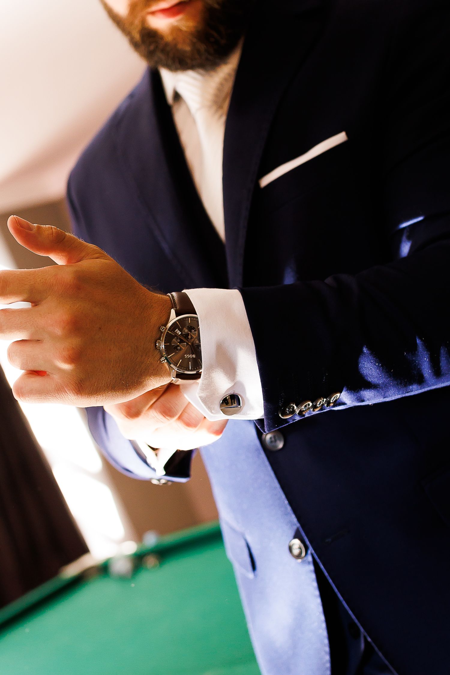 Elegant bearded groom in navy blue suit adjusting luxury watch cufflinks before wedding ceremony.