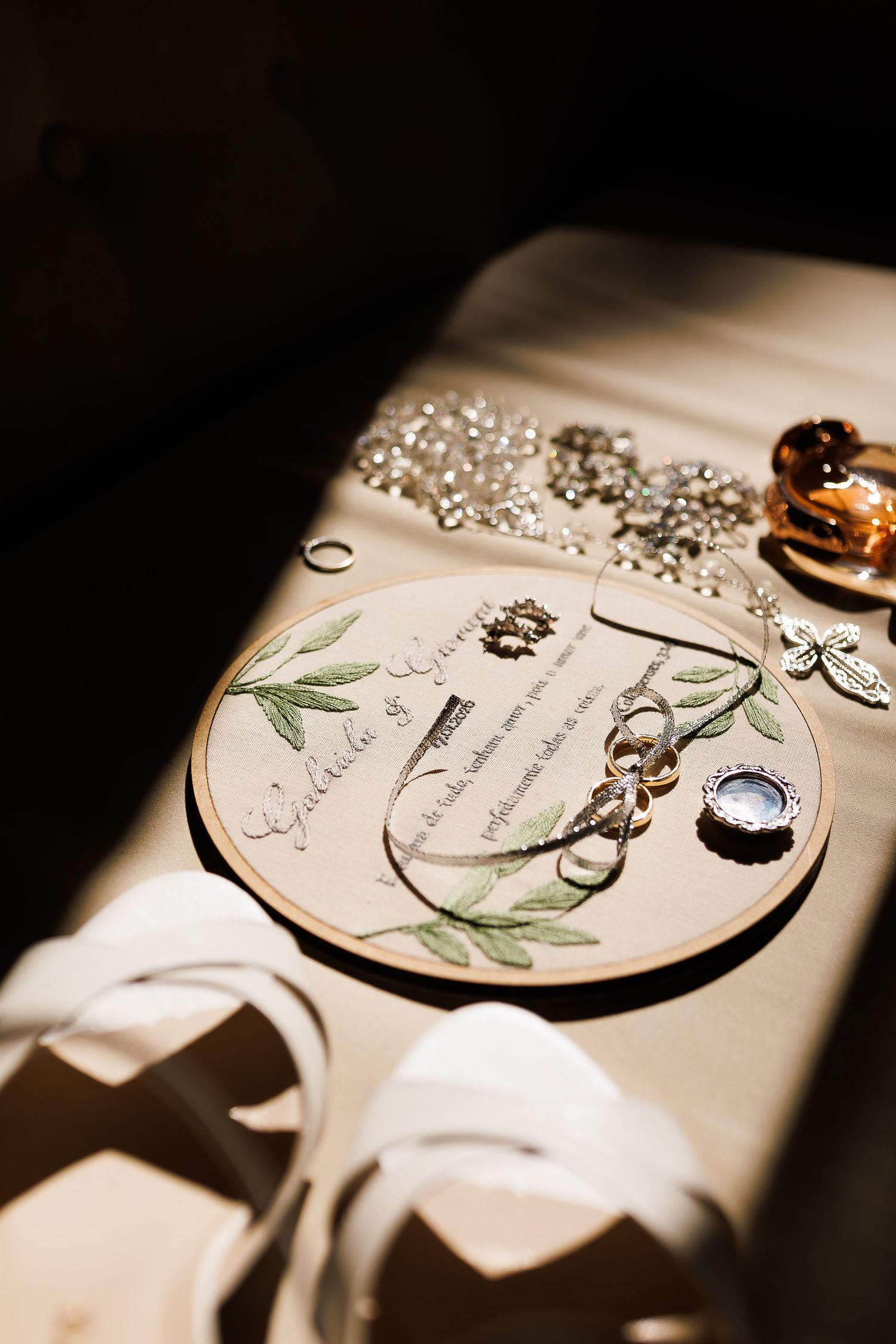 Wedding flat lay with floral embroidery hoop, gold rings, silver jewelry, and elegant invitation on cream fabric.