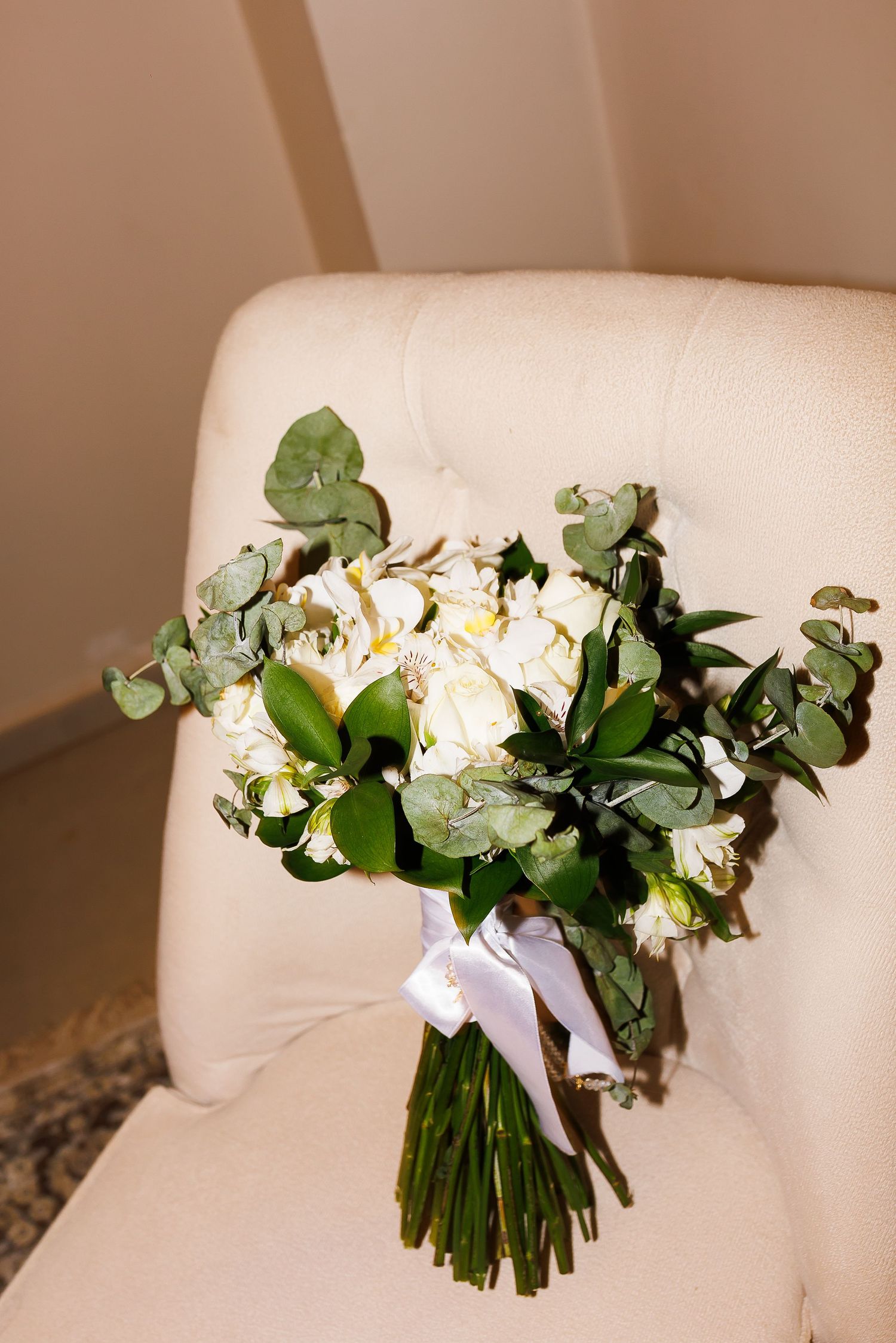 Elegant bridal bouquet of white peonies and eucalyptus tied with white ribbon, resting on a cream upholstered chair.