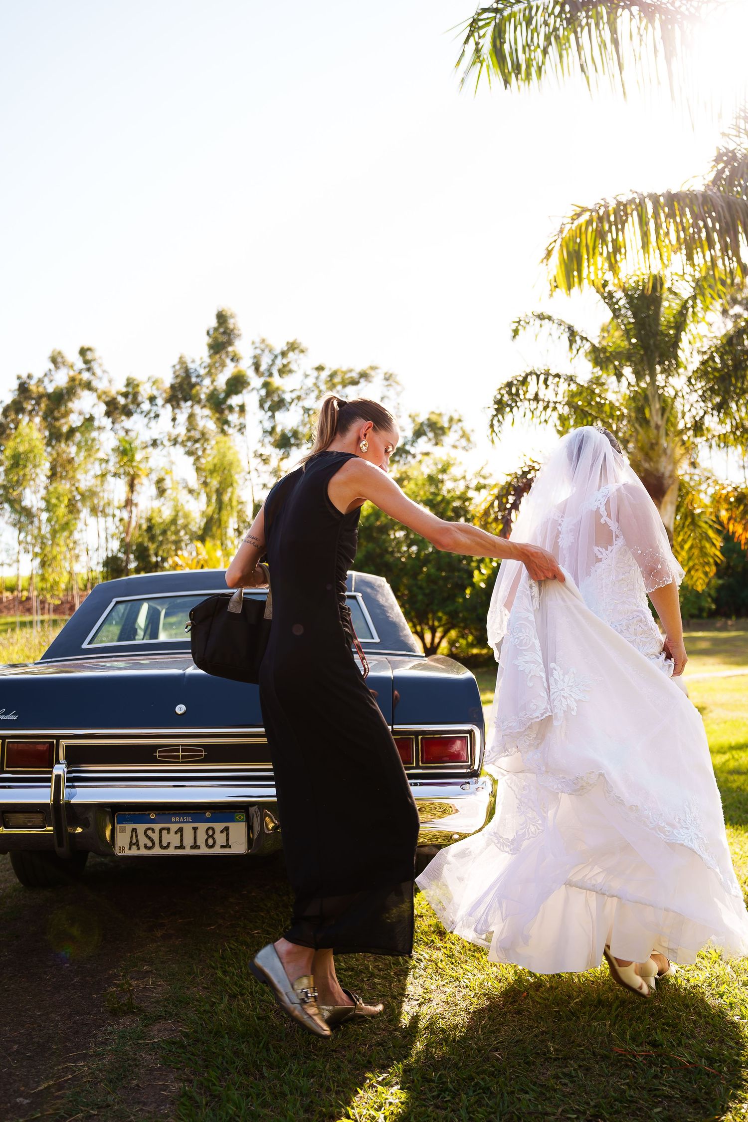 Bride in white gown and veil assisted by woman in black dress near vintage car outdoors on sunny day.