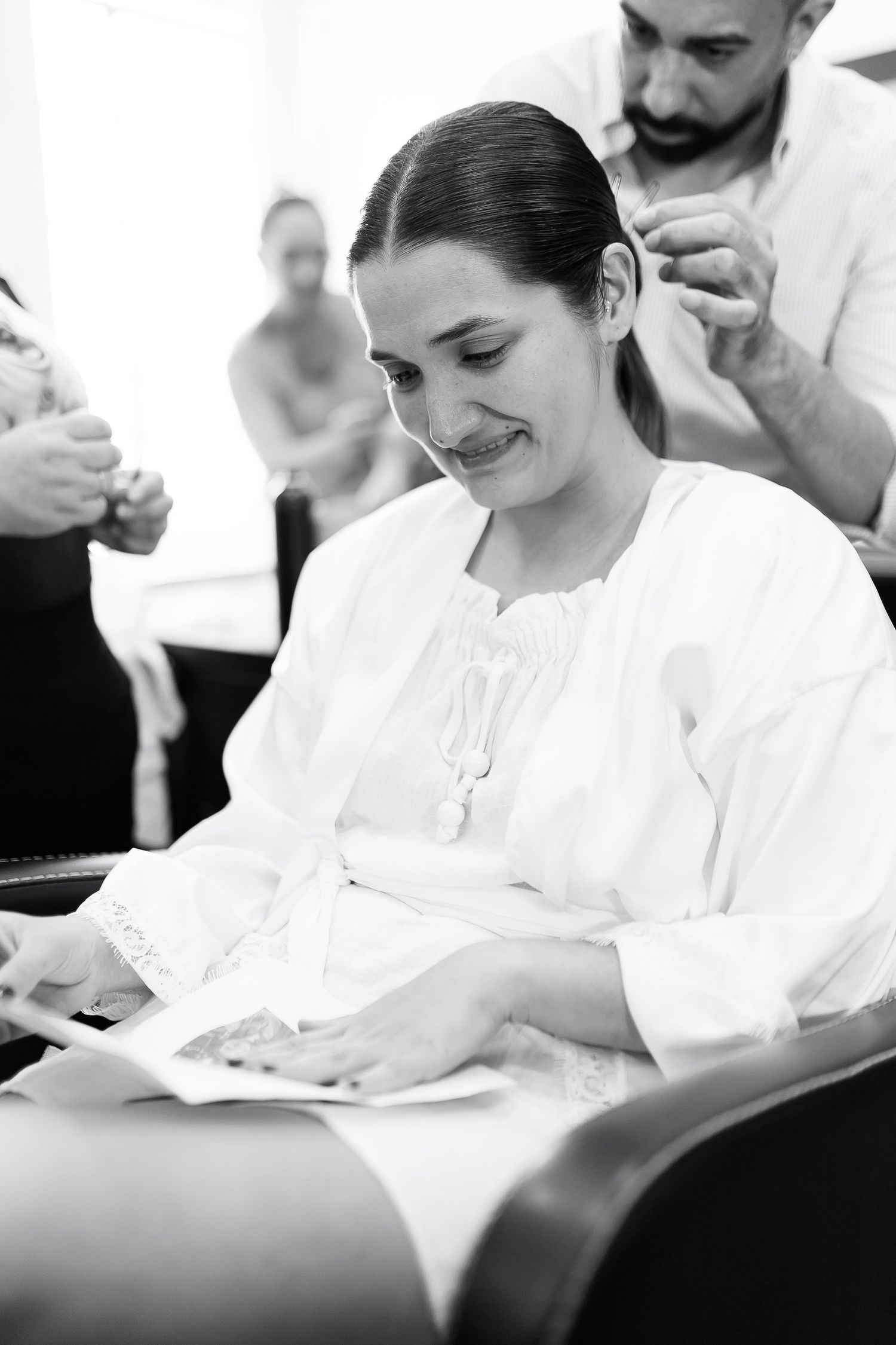Black and white photo of a smiling woman in white blouse reading, while a hairdresser works behind her.