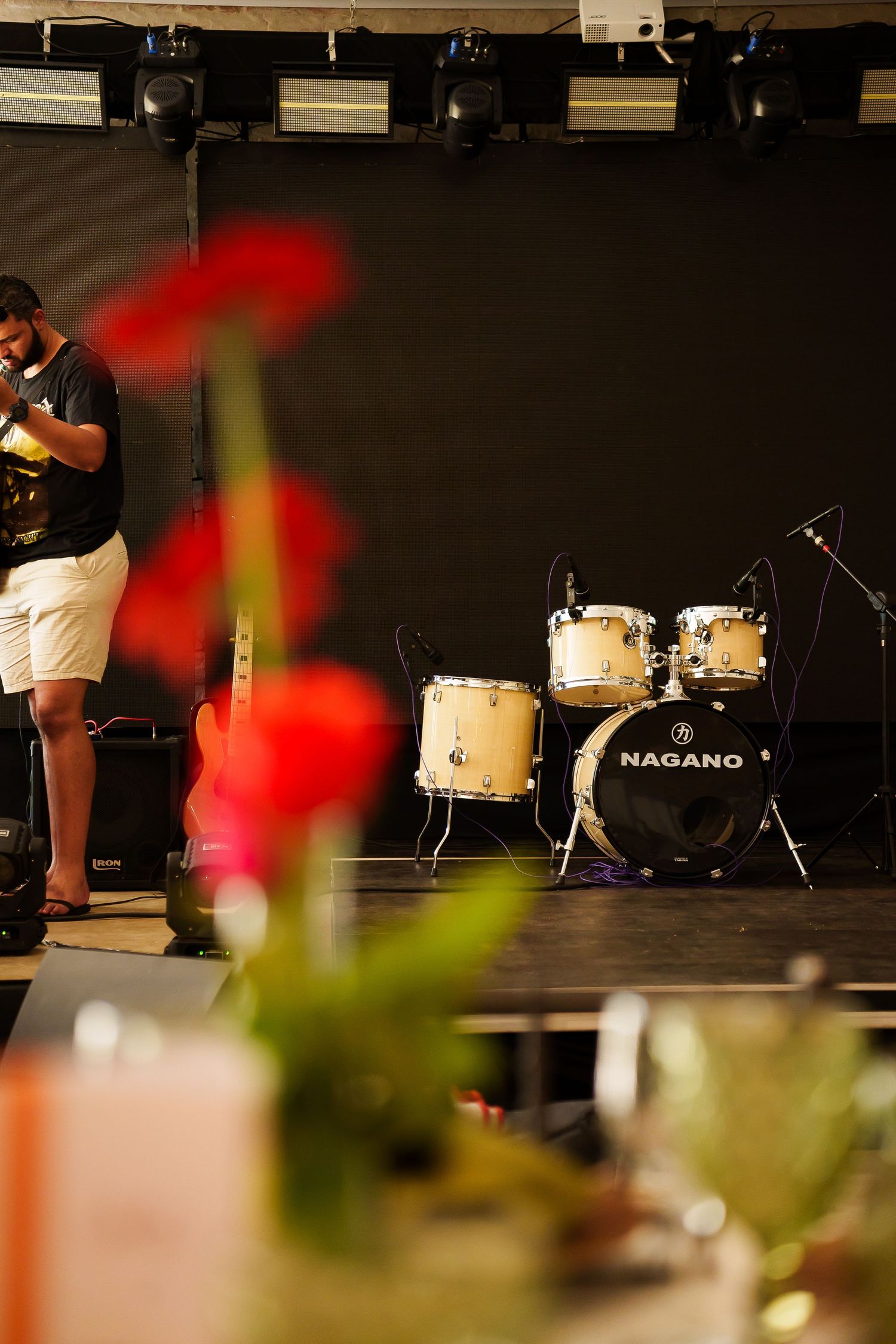 Musician on stage with Nagano drum kit, red flowers in foreground at live music venue.