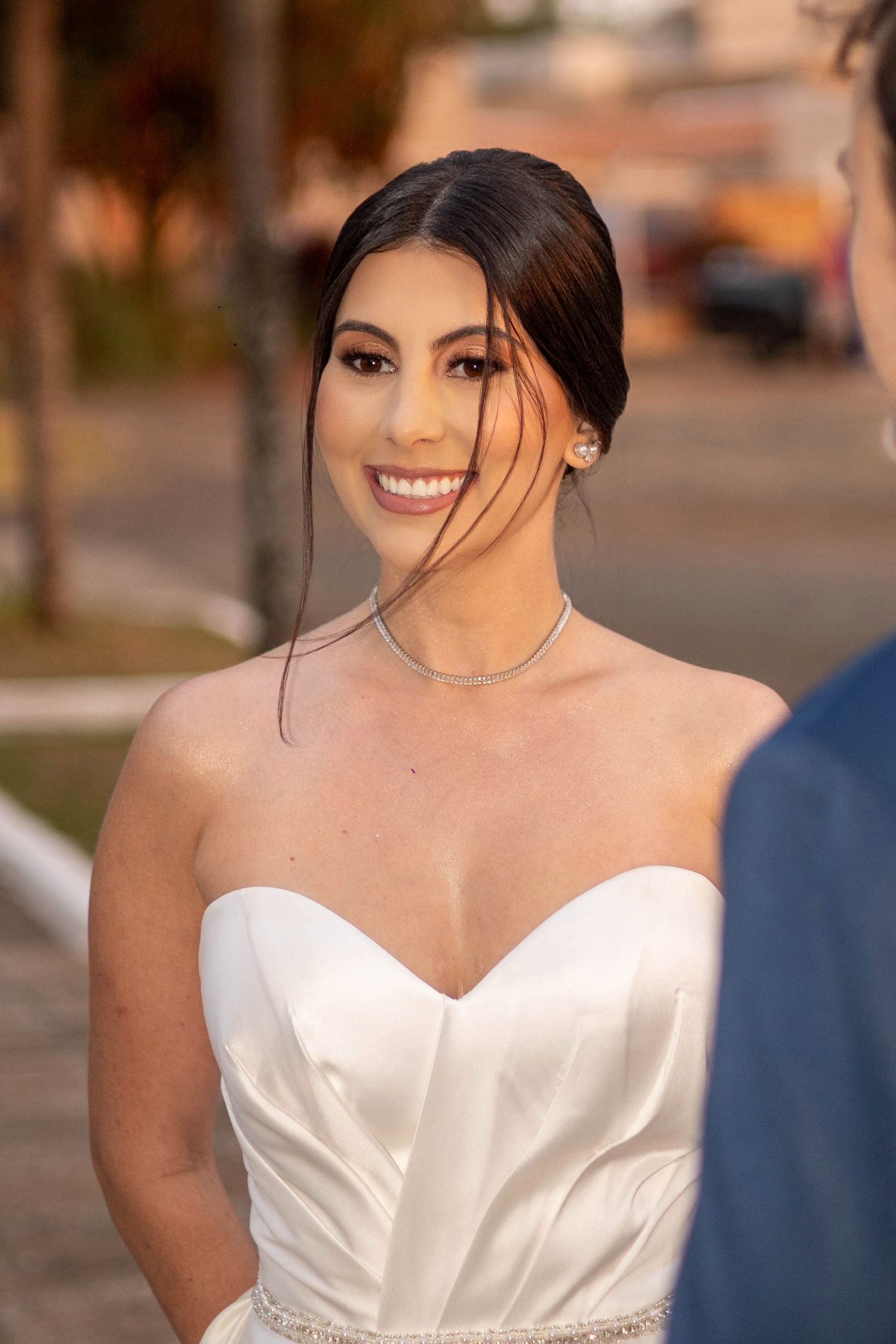 Smiling bride in a white strapless satin gown with diamond necklace and earrings, posing outdoors at golden hour.