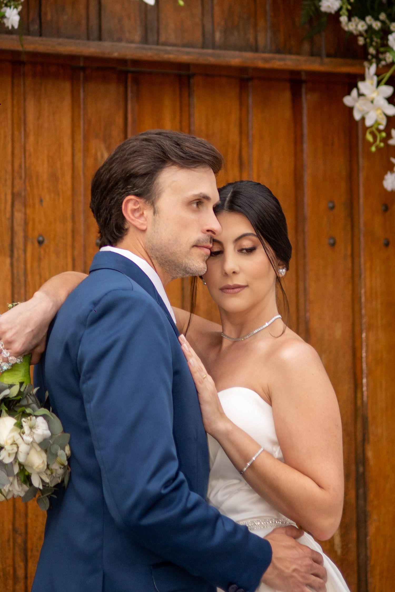 Bride and groom embracing tenderly in front of rustic wooden doors, bride in strapless gown holding bouquet of white flowers.