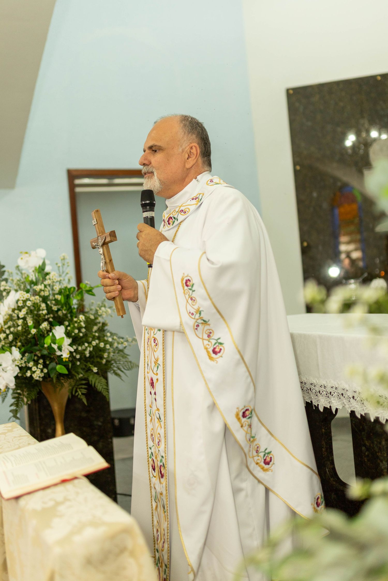 Catholic priest in white vestments holds a crucifix and microphone while delivering a sermon at an altar.