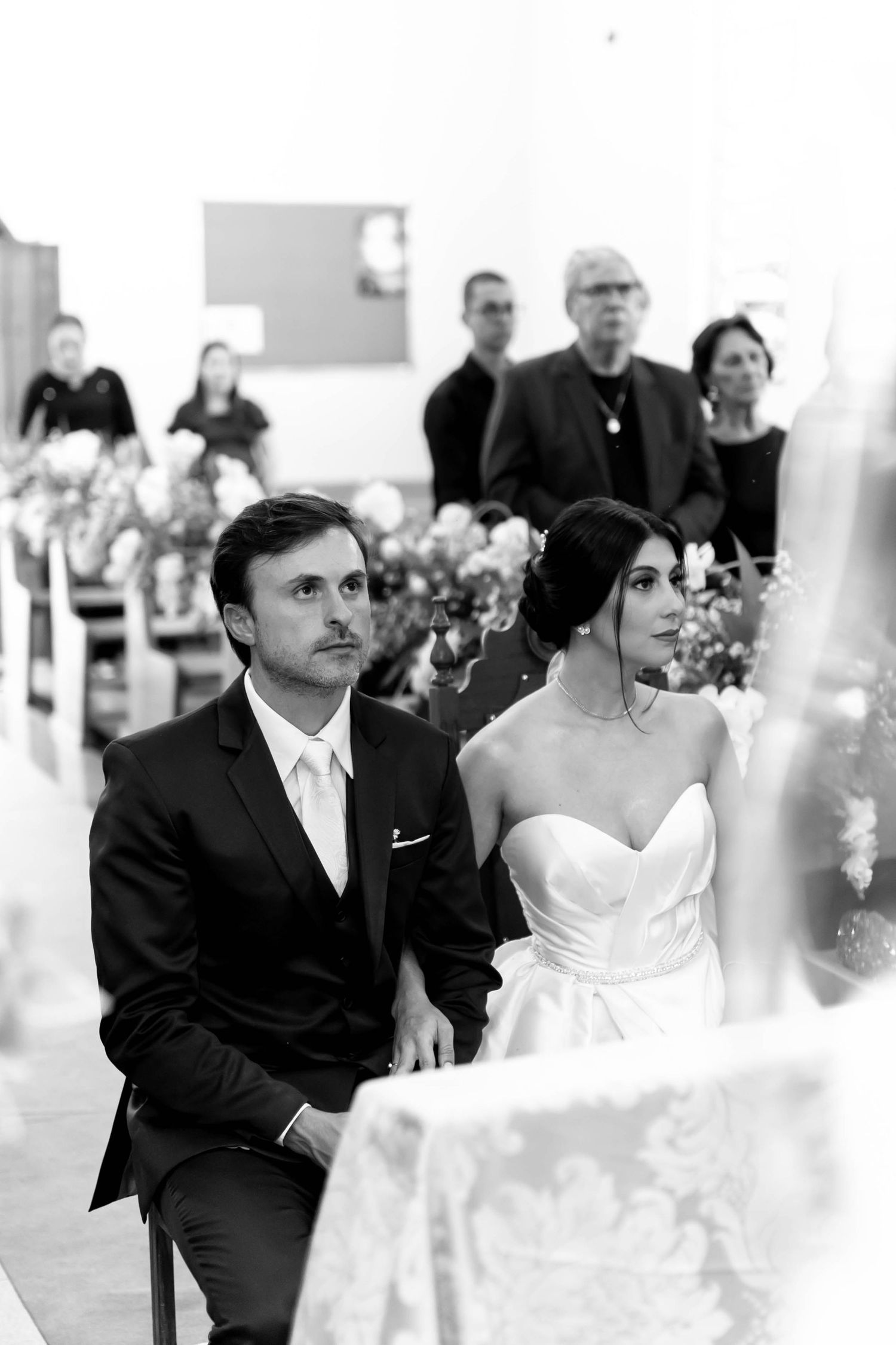 Black and white photo of a bride and groom sitting at the altar during their wedding ceremony in a church.
