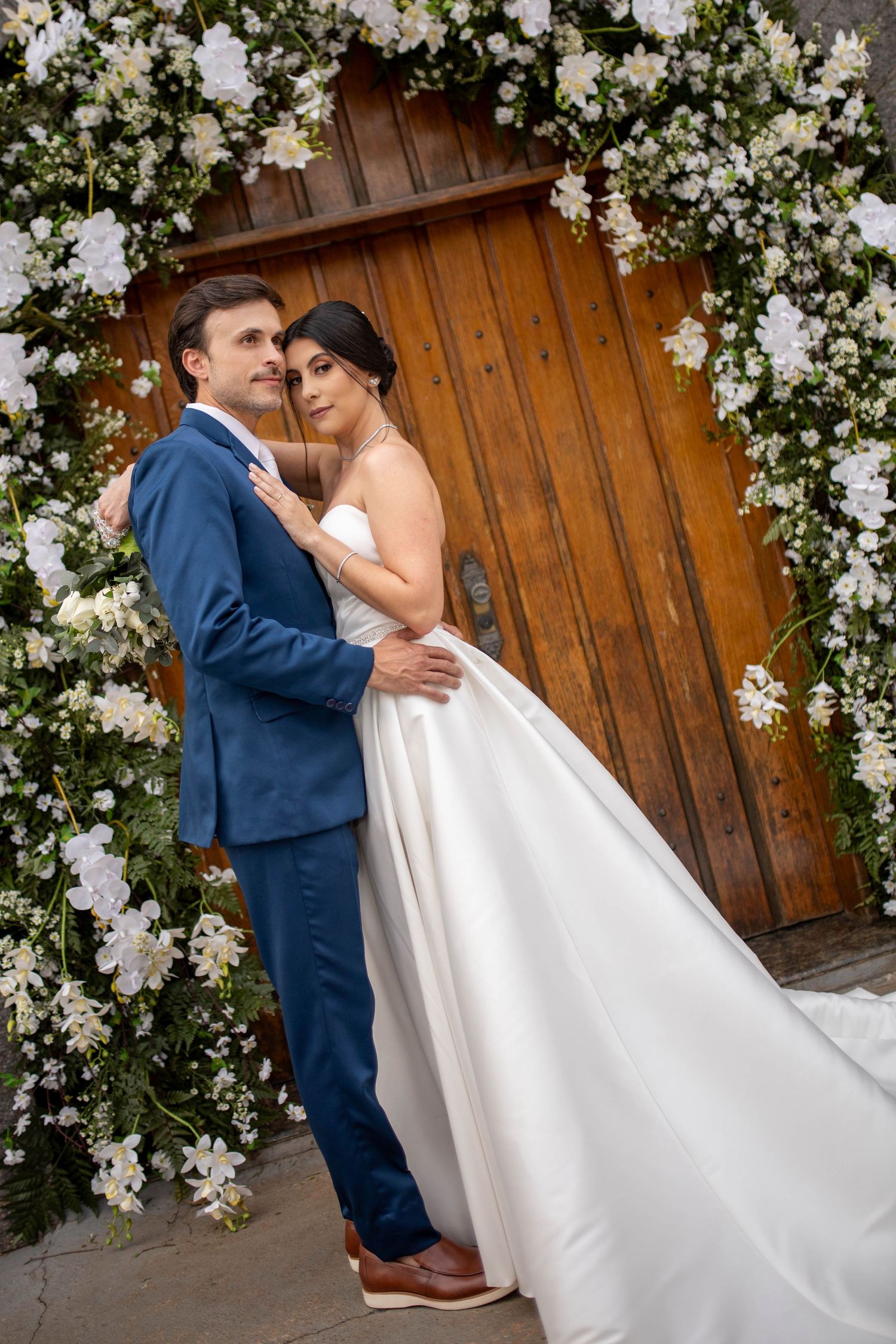 Bride and groom embrace before floral arch with white flowers and wooden door on their wedding day.