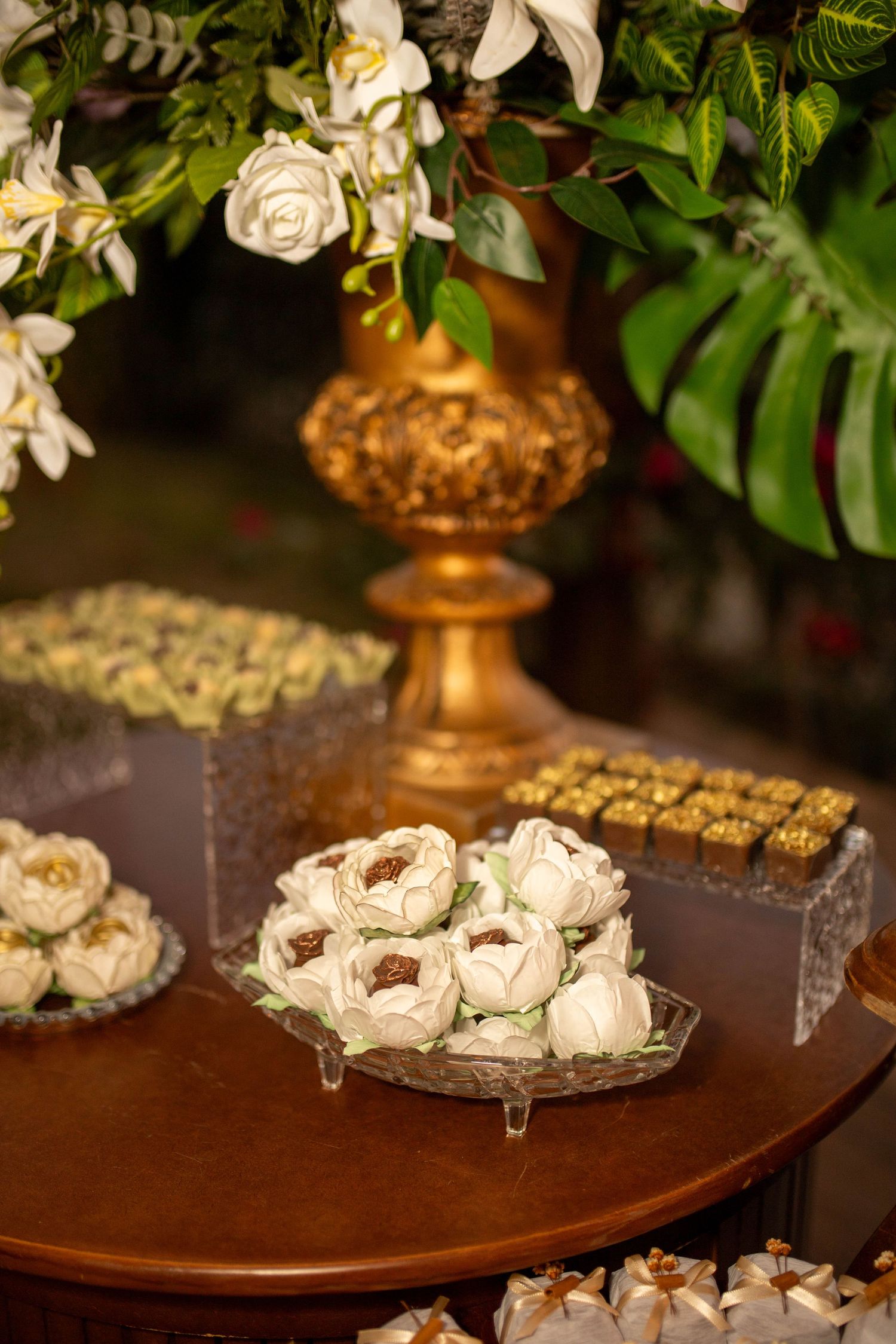Elegant dessert table with white chocolate flower candies on crystal plate, gold vase with white roses, and assorted sweets.