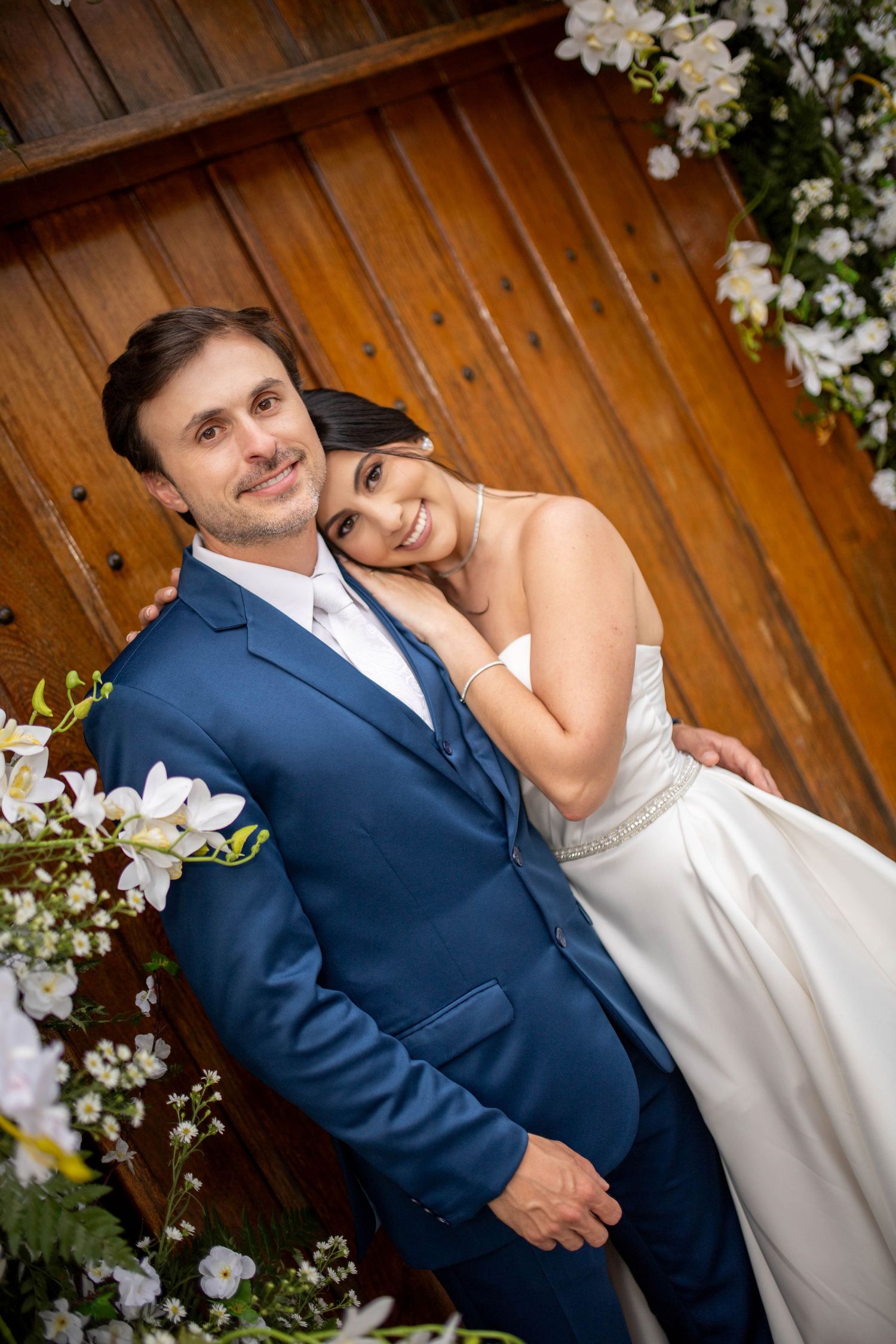 A smiling bride in a white gown leans on her groom in a blue suit, posing together near a wooden door with white flowers.
