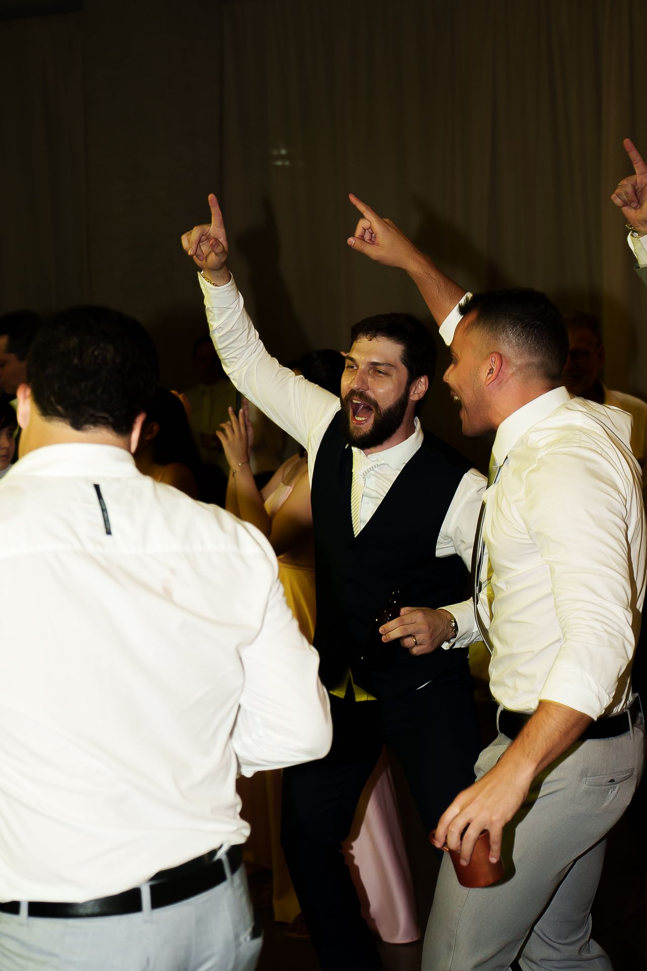Men in white shirts and a vest dance energetically at a wedding reception, pointing up and celebrating.