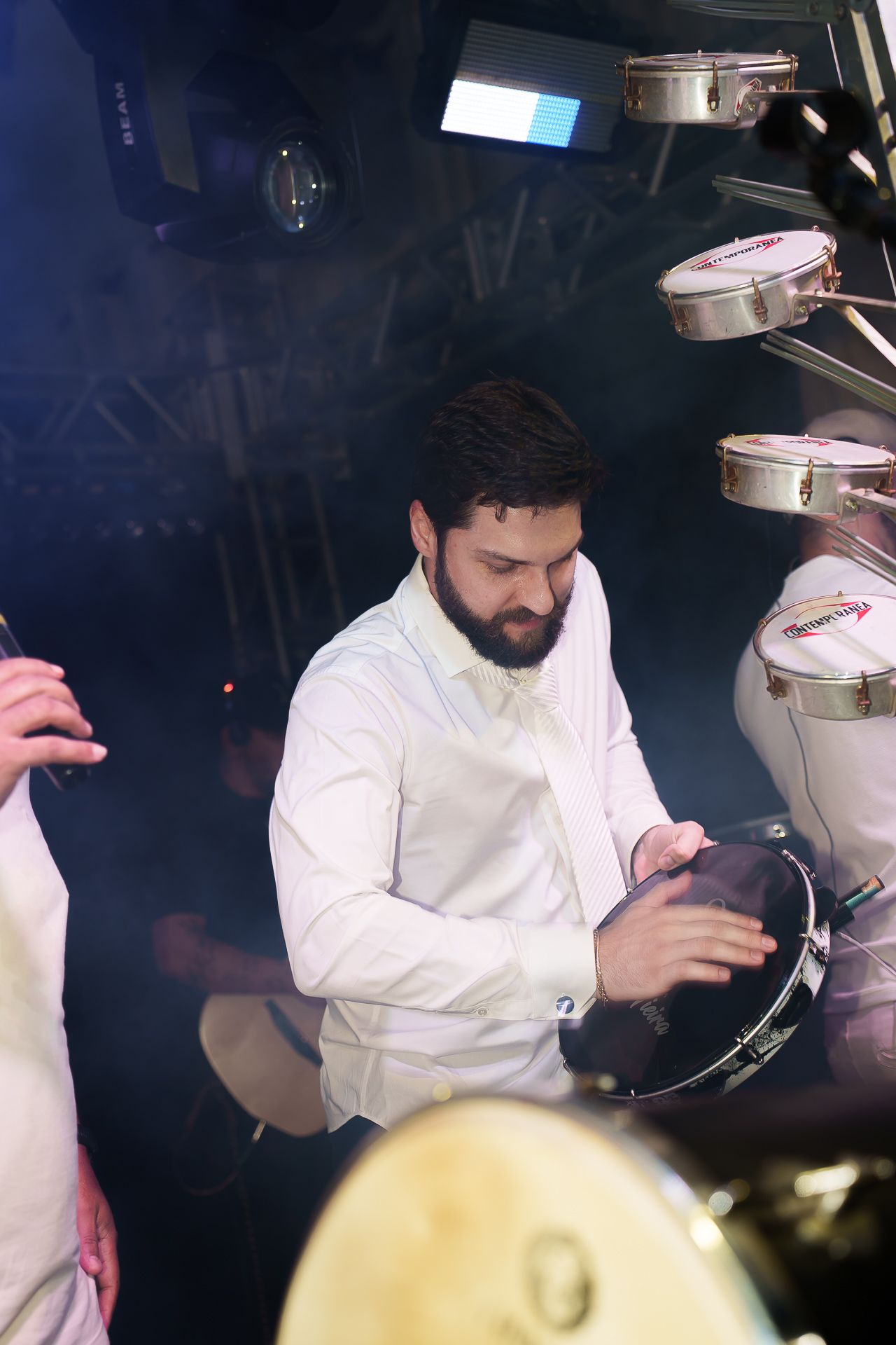 Bearded man in white shirt playing a tambourine surrounded by drums in a smoky nightclub setting.