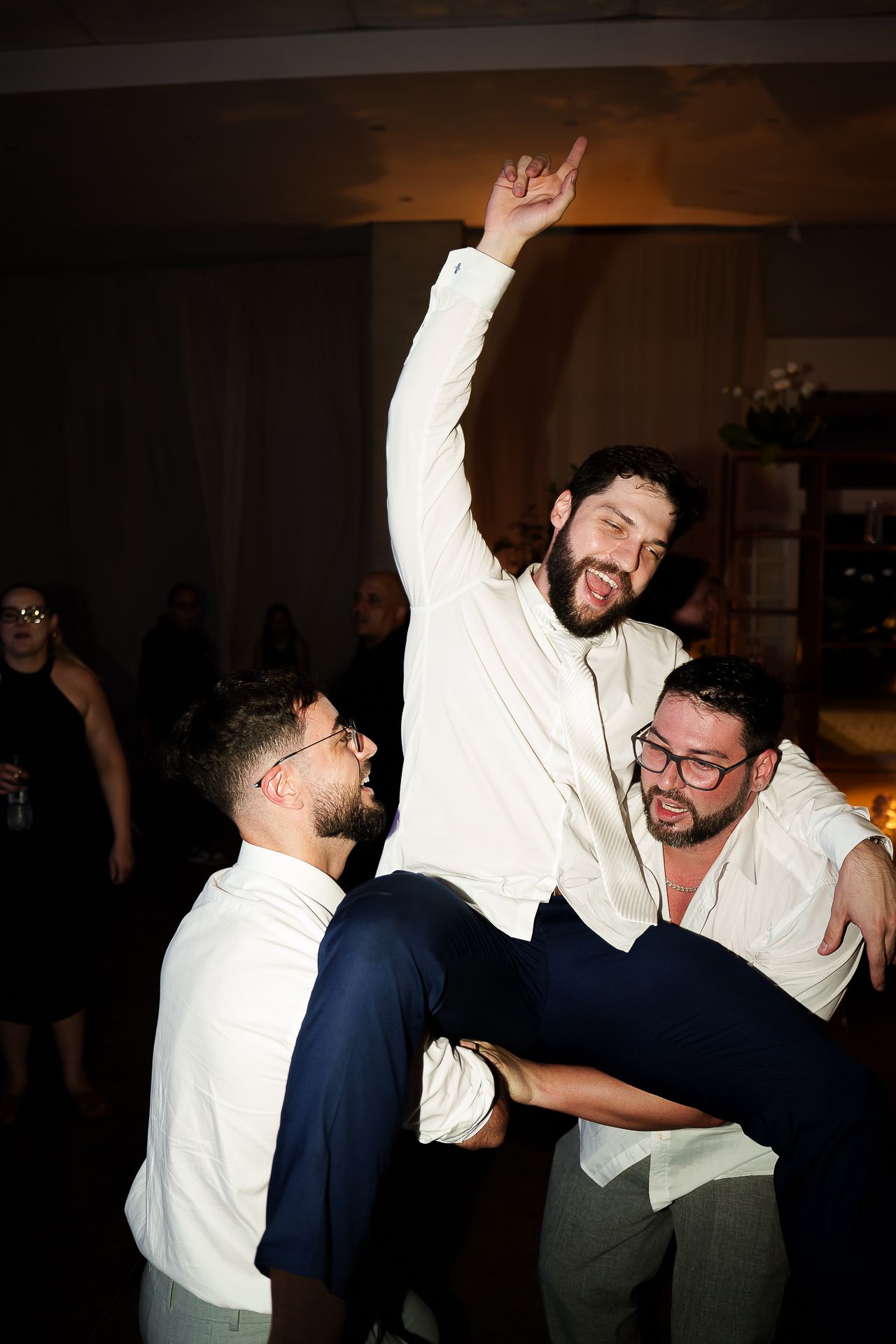 Two groomsmen lift a bearded man in a white shirt at a wedding reception celebration.