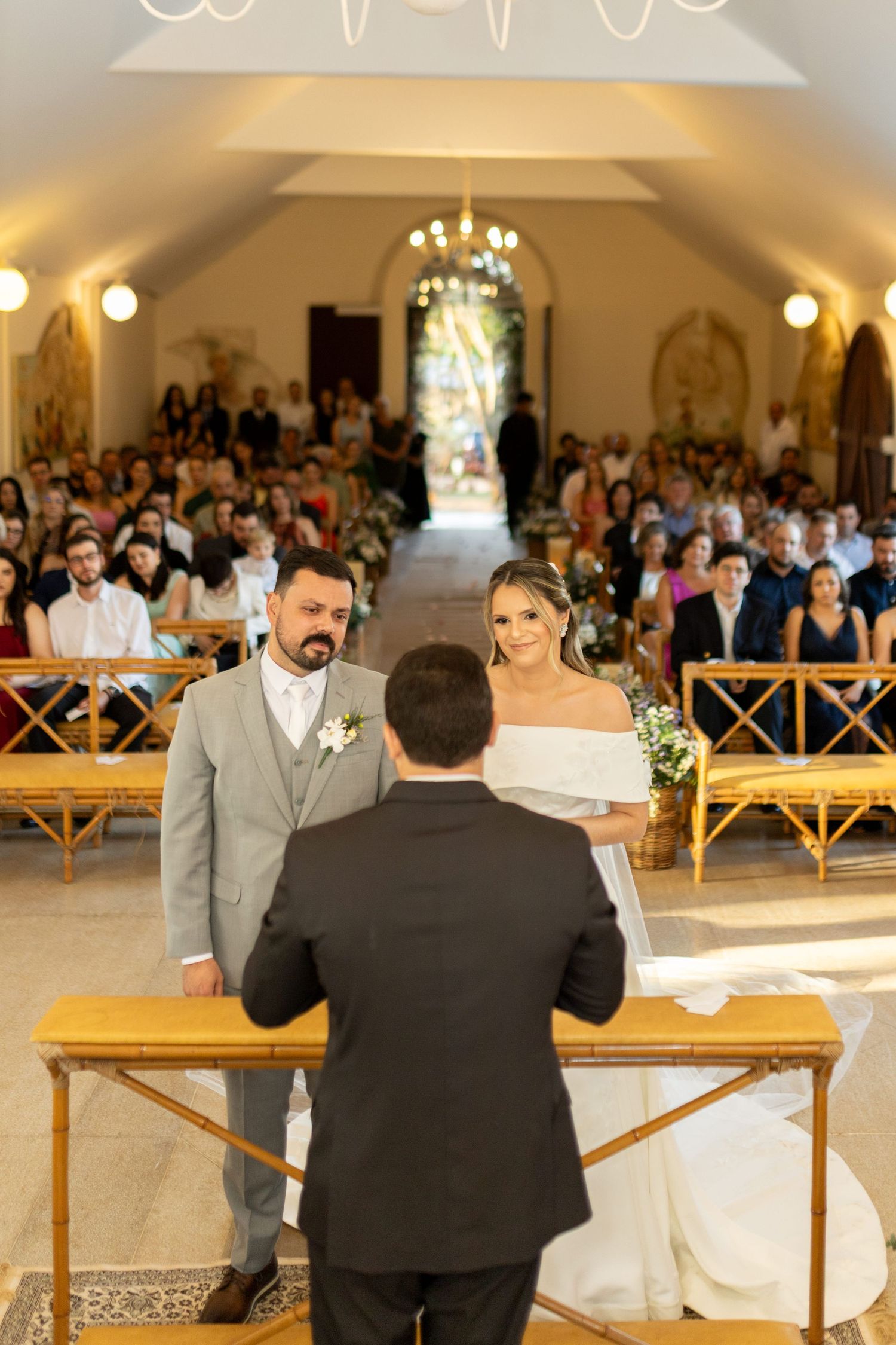 Bride and groom exchange vows at the altar in a sunlit chapel filled with seated wedding guests.