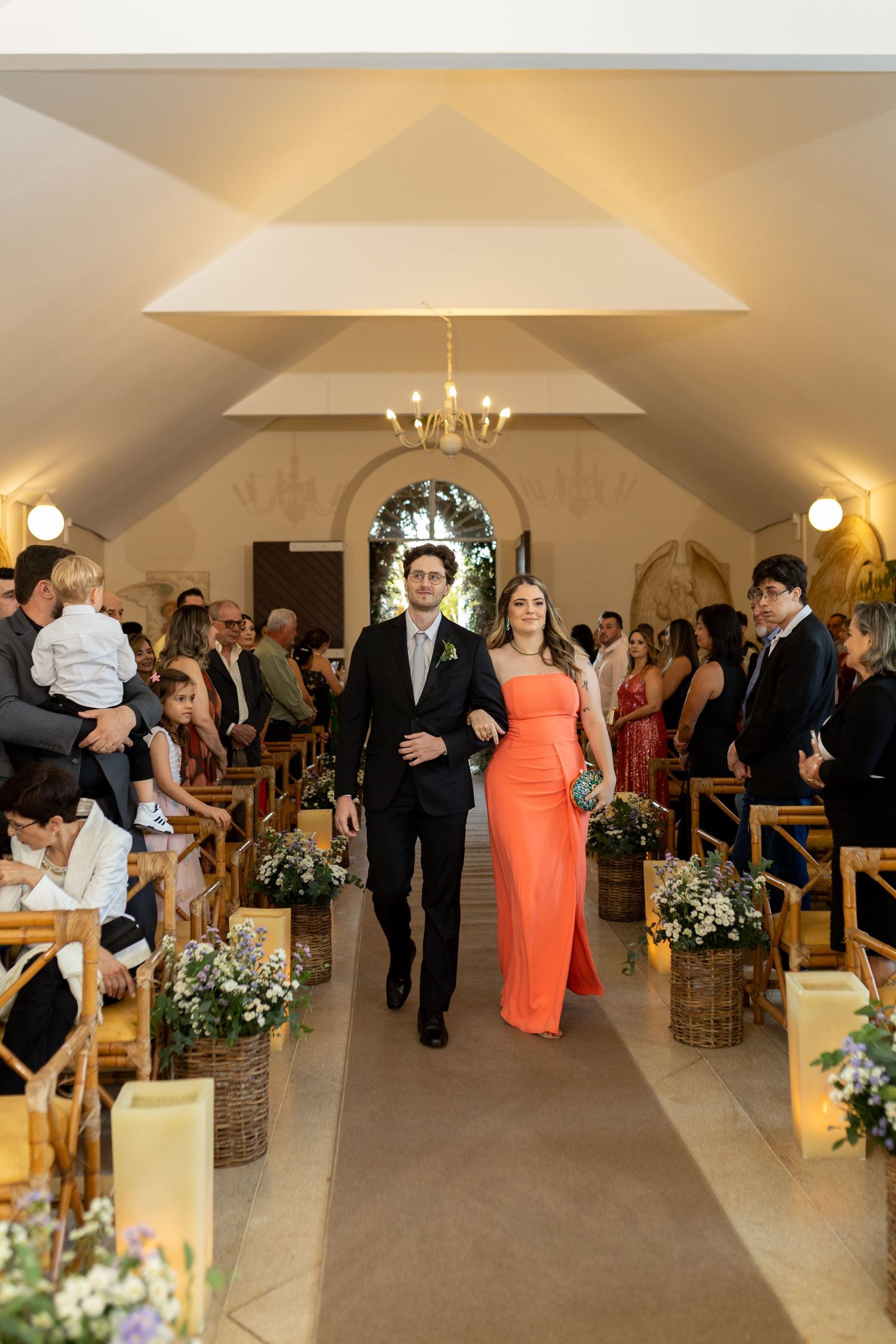 A couple walks down the aisle of a decorated chapel during a wedding ceremony, guests seated on wooden pews.