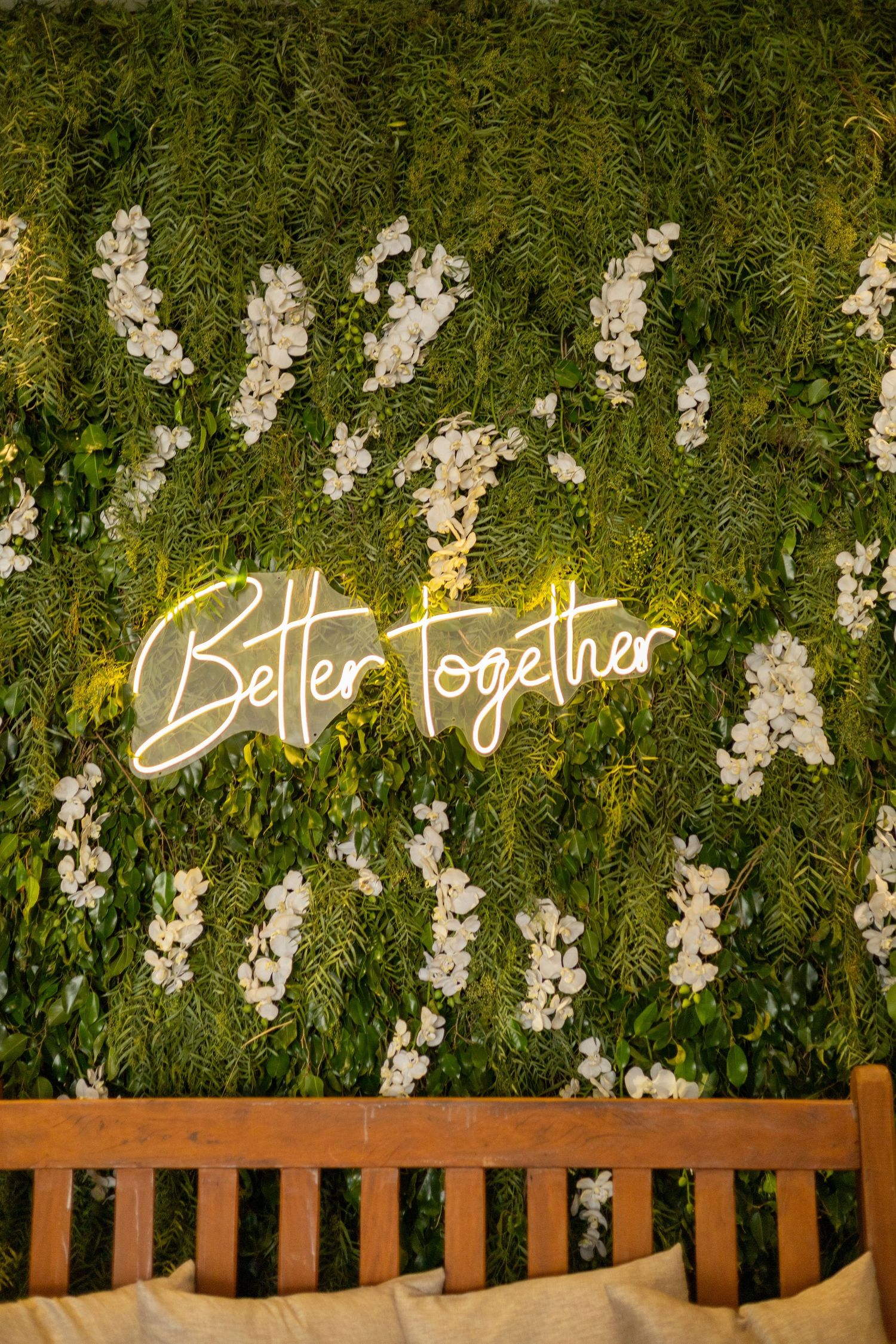 Neon 'Better Together' sign glowing on a lush green floral wall with white orchids above a wooden bench.