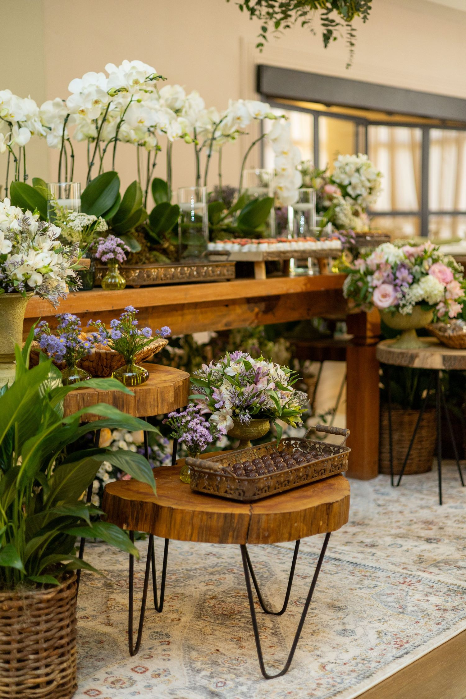 Elegant floral display with white orchids, mixed blooms on rustic wooden tables with hairpin legs on a patterned rug.