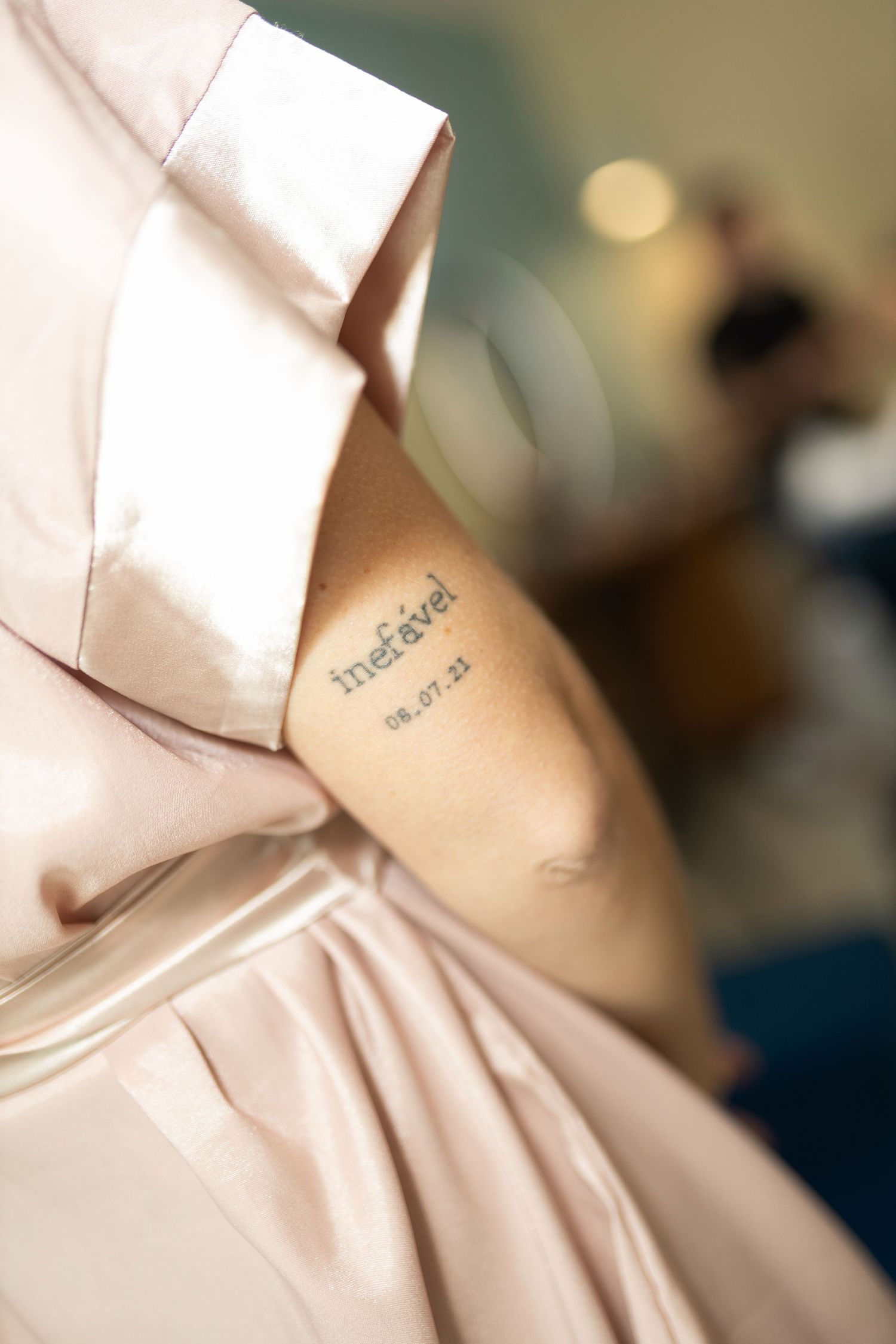 Close-up of a tattoo reading 'inefável' and date 06.07.21 on an arm, beside a pink satin wedding dress.