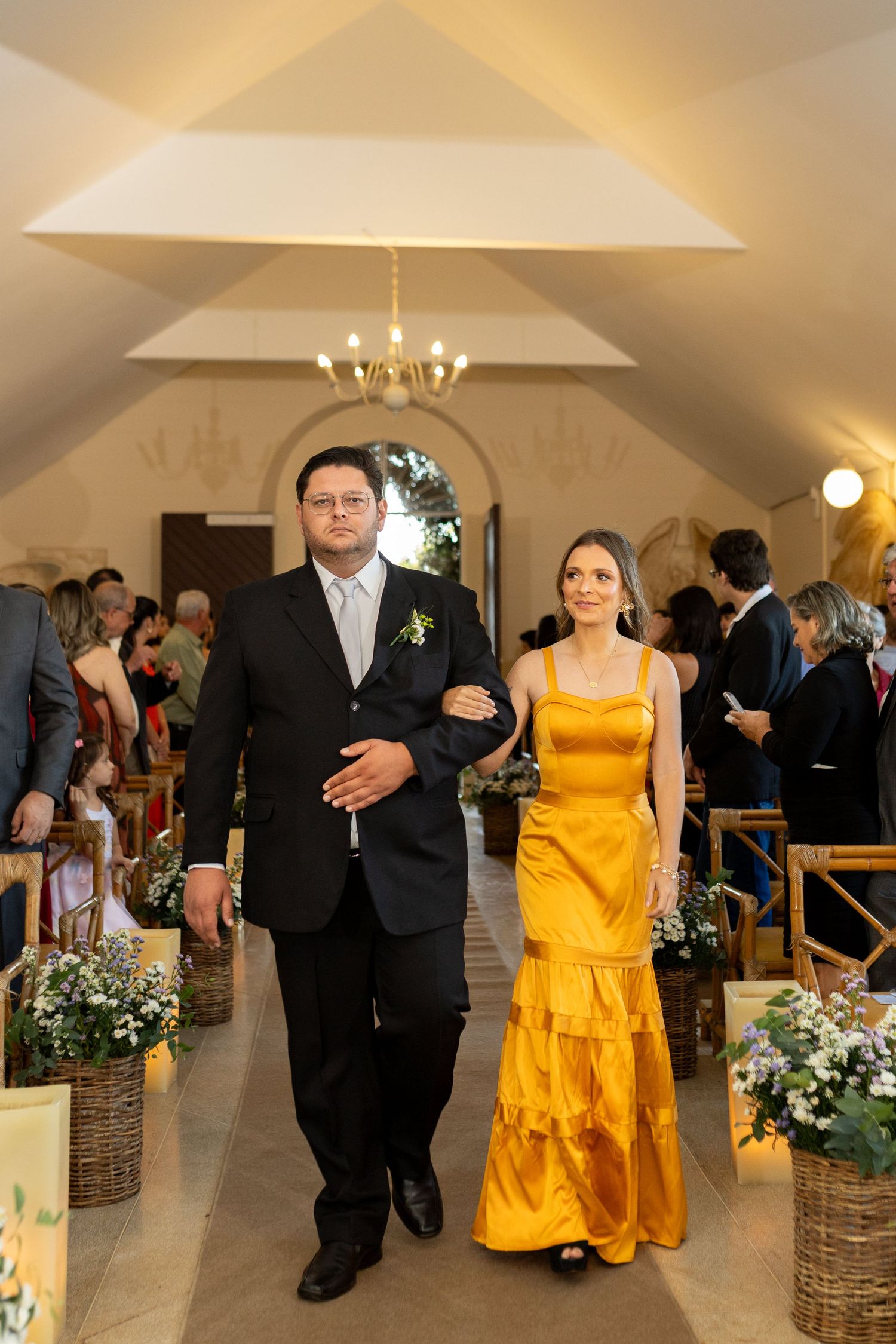 Couple walking down wedding aisle, man in black suit, woman in golden yellow gown, floral decorations.