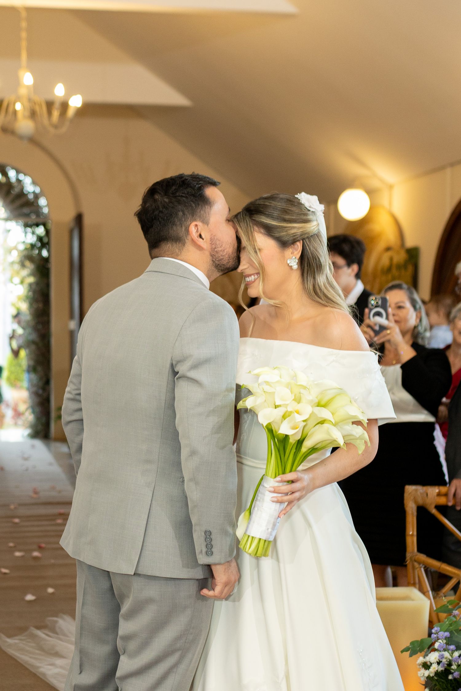 Bride and groom share a kiss at wedding ceremony, bride holding white calla lily bouquet in elegant off-shoulder gown.