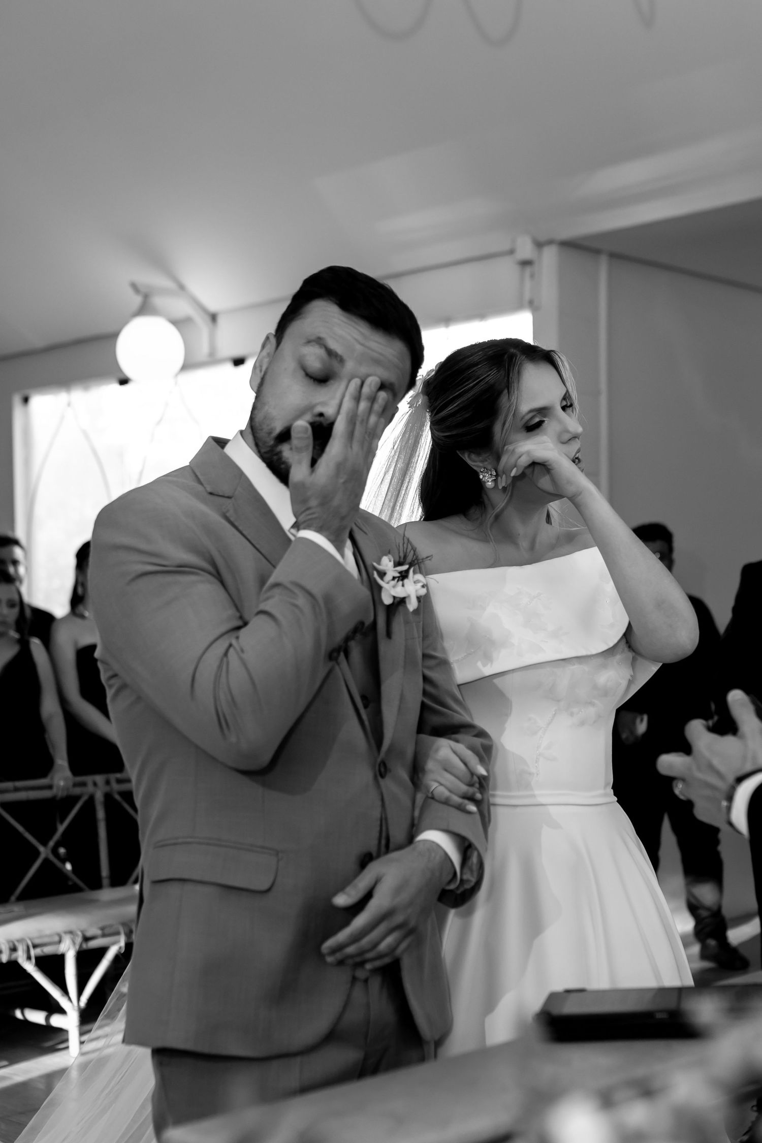 Black and white photo of emotional bride and groom wiping tears during wedding ceremony.