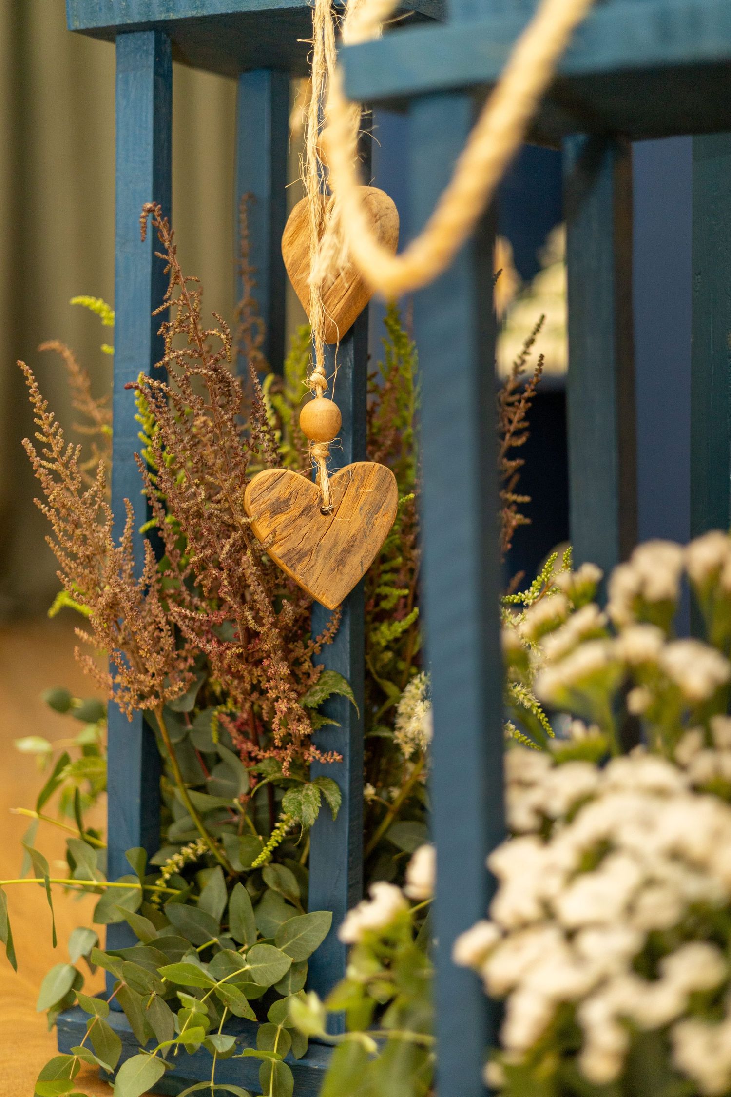 Wooden heart ornaments hanging on a blue wooden fence surrounded by dried flowers and green eucalyptus foliage.