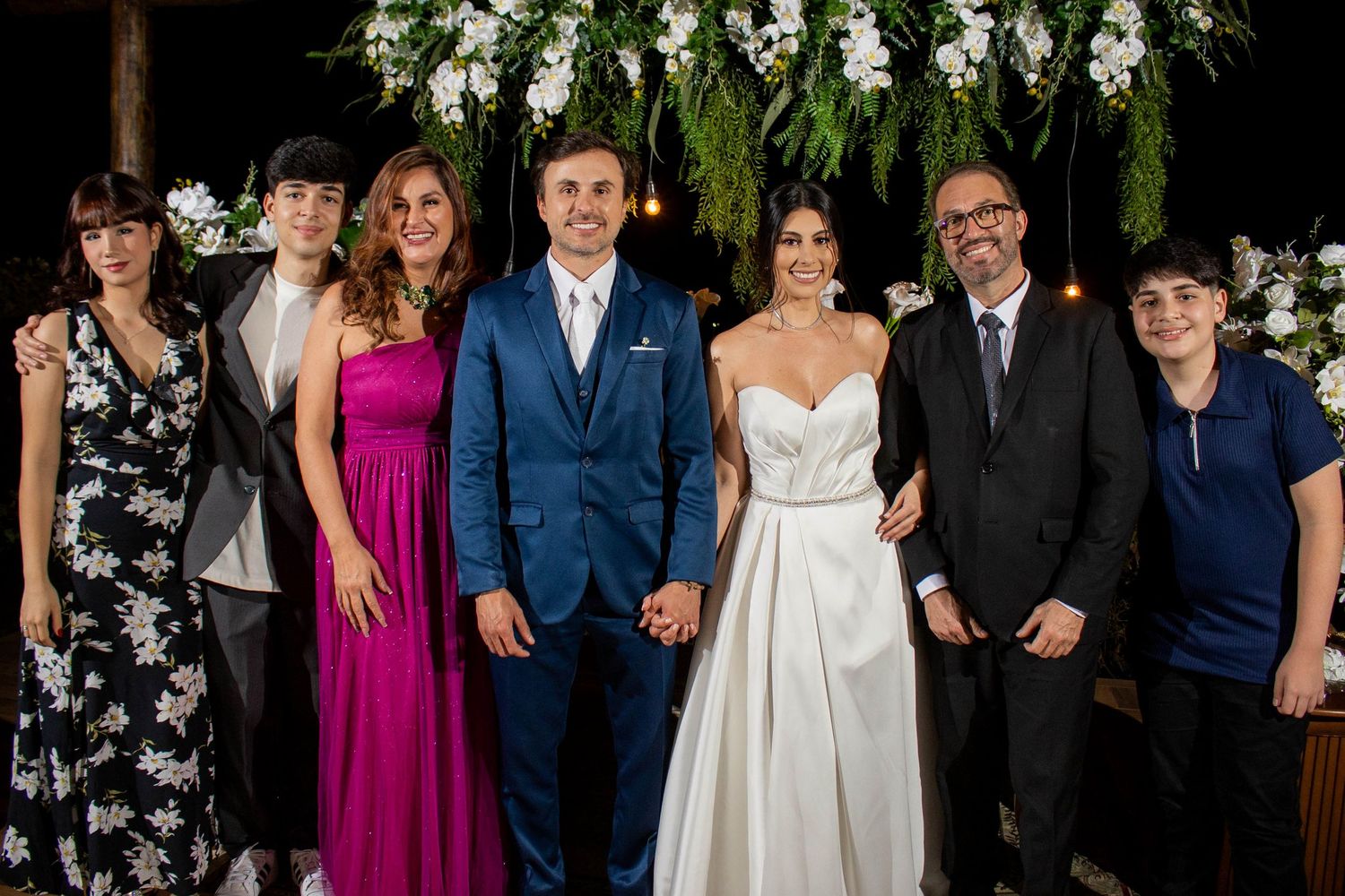 Wedding party posing together under floral arch, bride in white gown and groom in blue suit with family members.