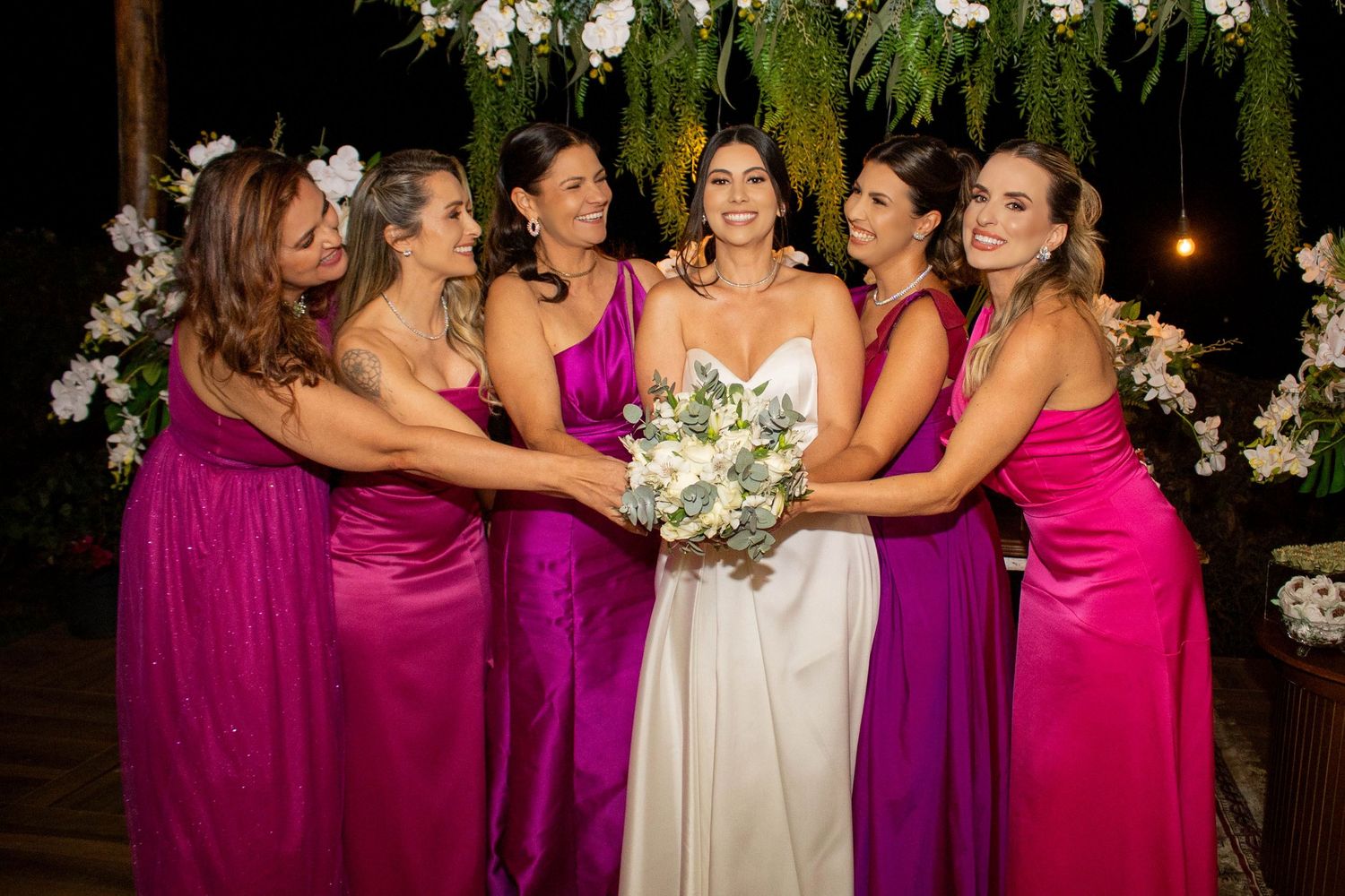 Smiling bride with white bouquet poses with five bridesmaids in magenta and purple gowns at evening wedding.