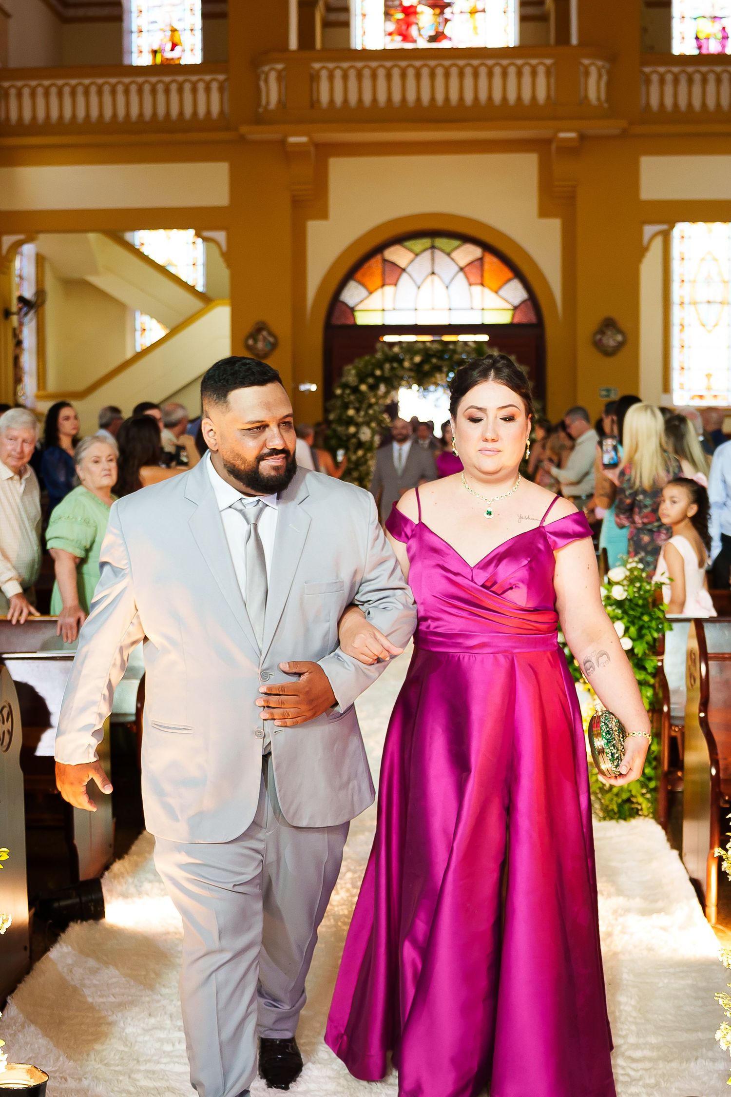 A man in a gray suit escorts a woman in a magenta satin gown down a church aisle during a wedding ceremony.