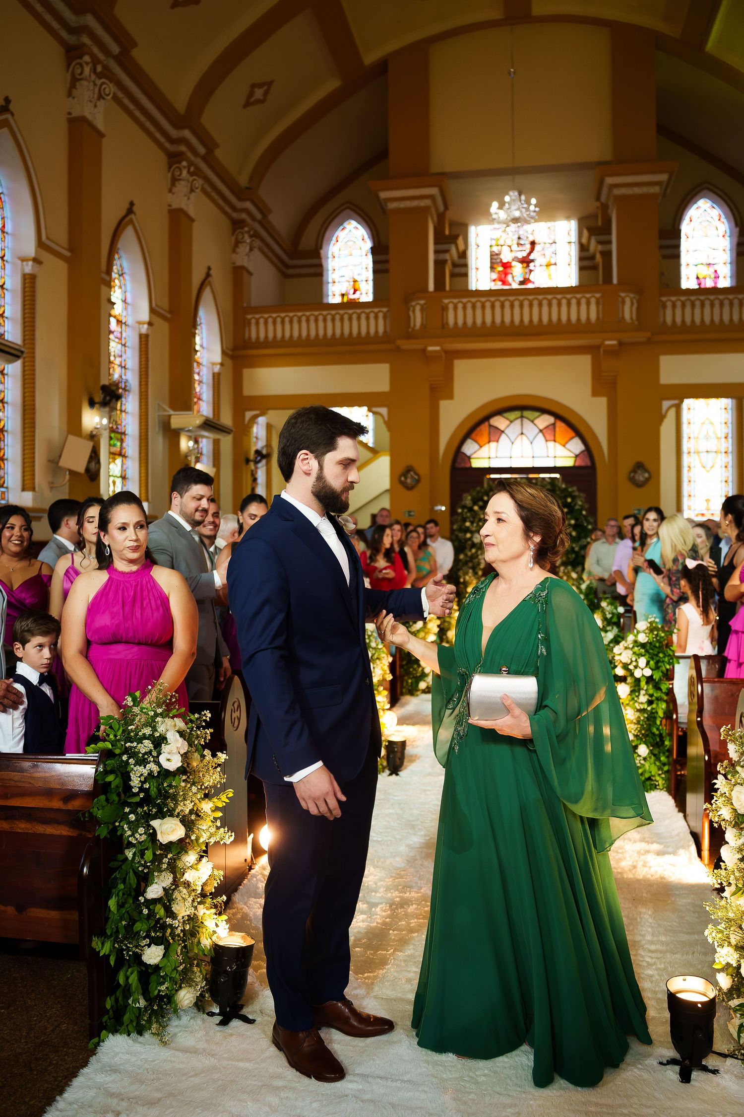 Groom in navy suit walks with woman in green gown down floral-decorated church aisle during wedding ceremony.