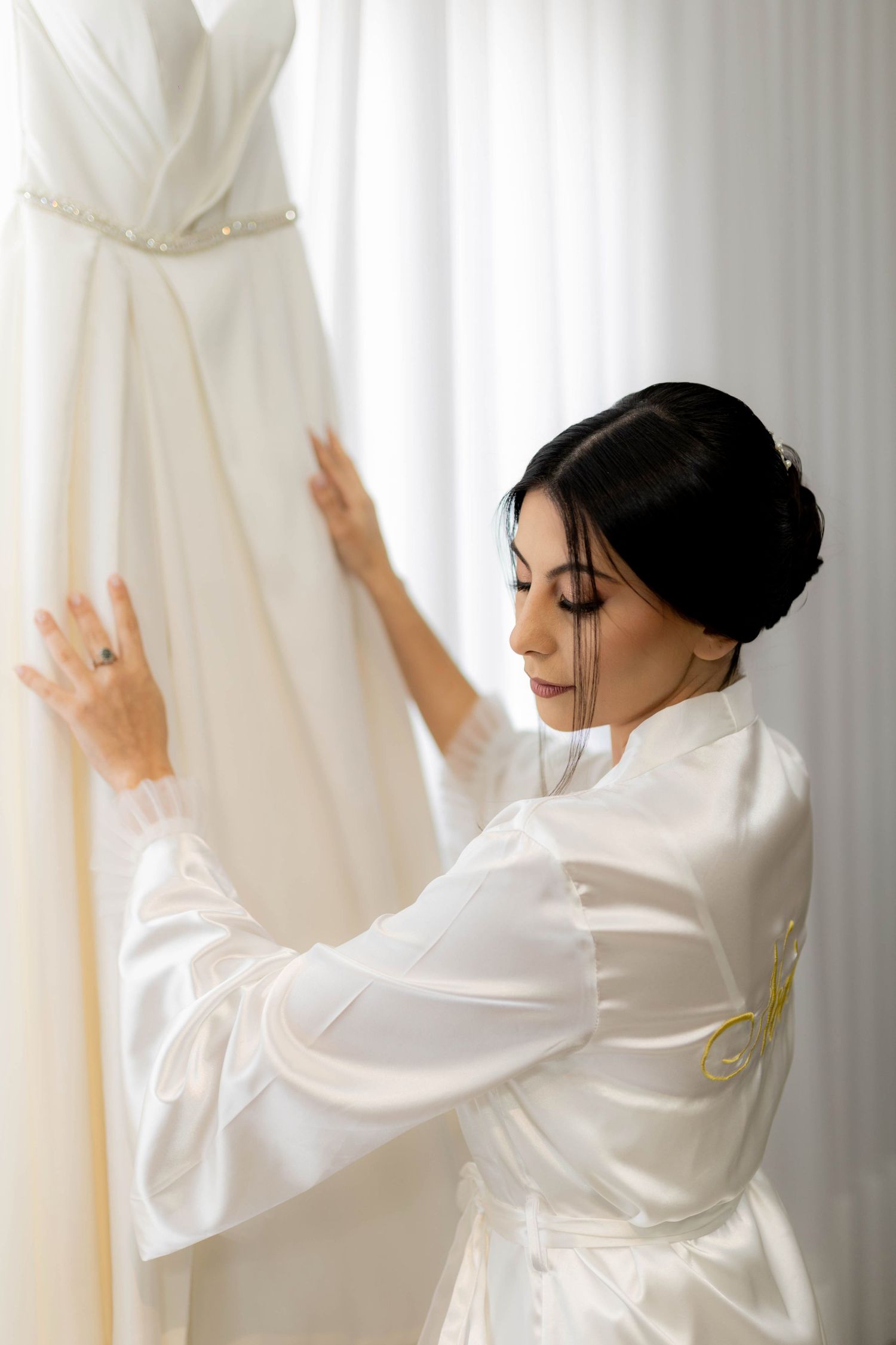 Bride in white satin robe admiring her elegant wedding dress hanging by a bright window before the ceremony.