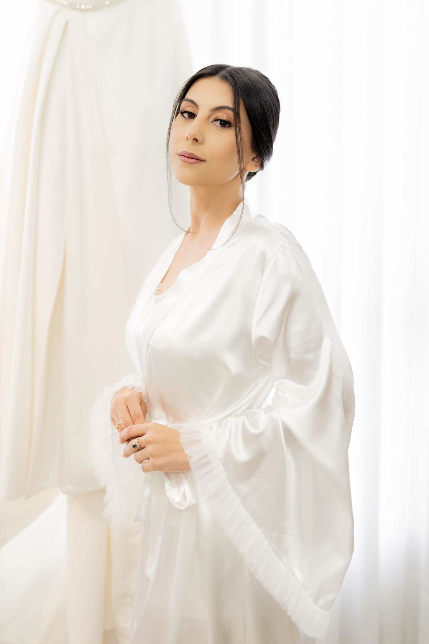 Bride in white satin robe preparing for wedding day, standing in front of bridal gown in bright room.