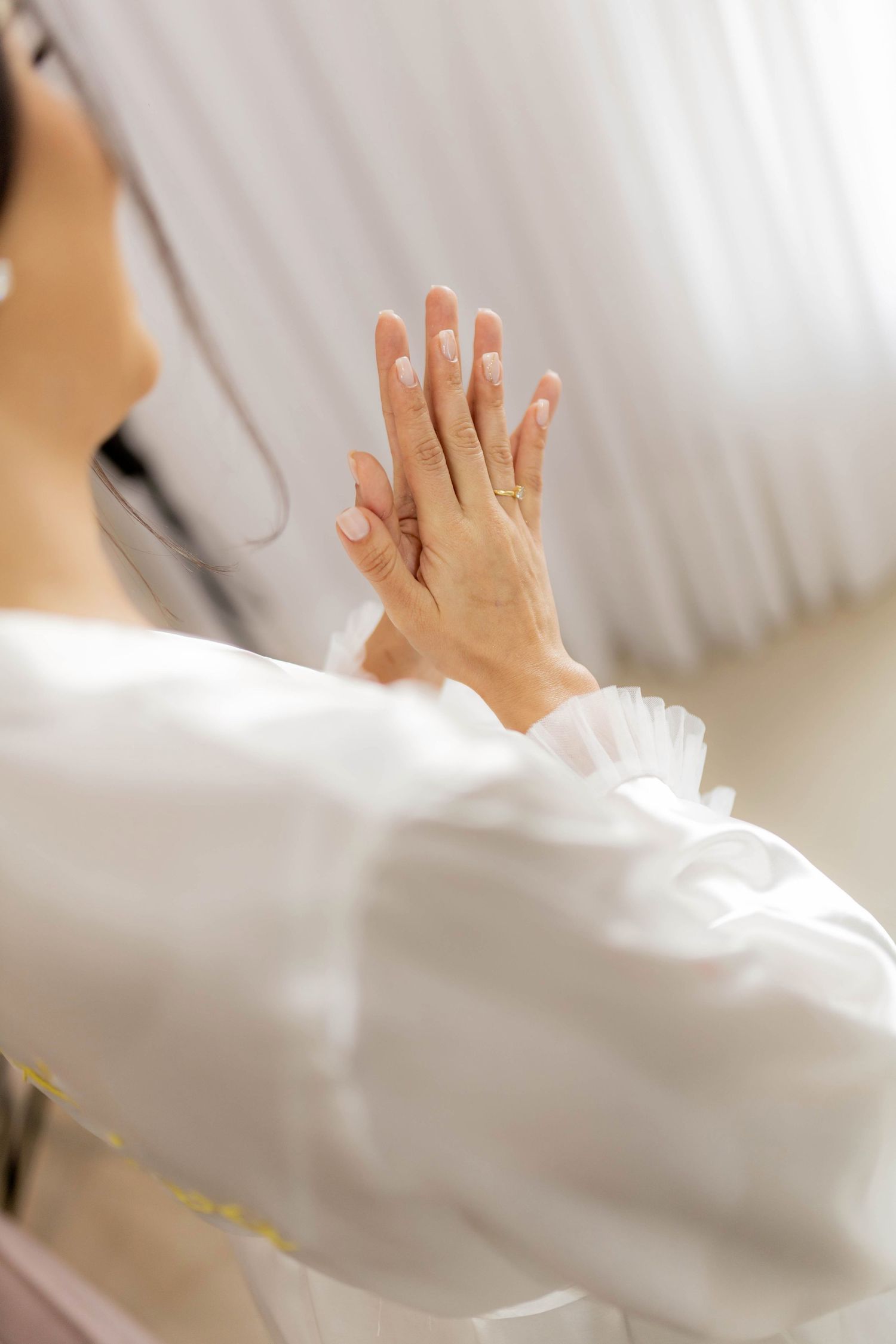 A bride in a white robe and veil admires her engagement ring while pressing her hands together before her wedding ceremony.