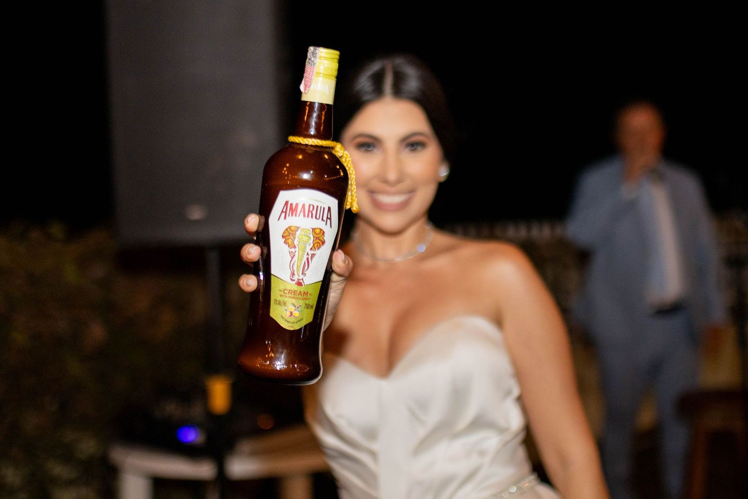 Woman in white dress holding up a bottle of Amarula cream liqueur at an outdoor evening event.
